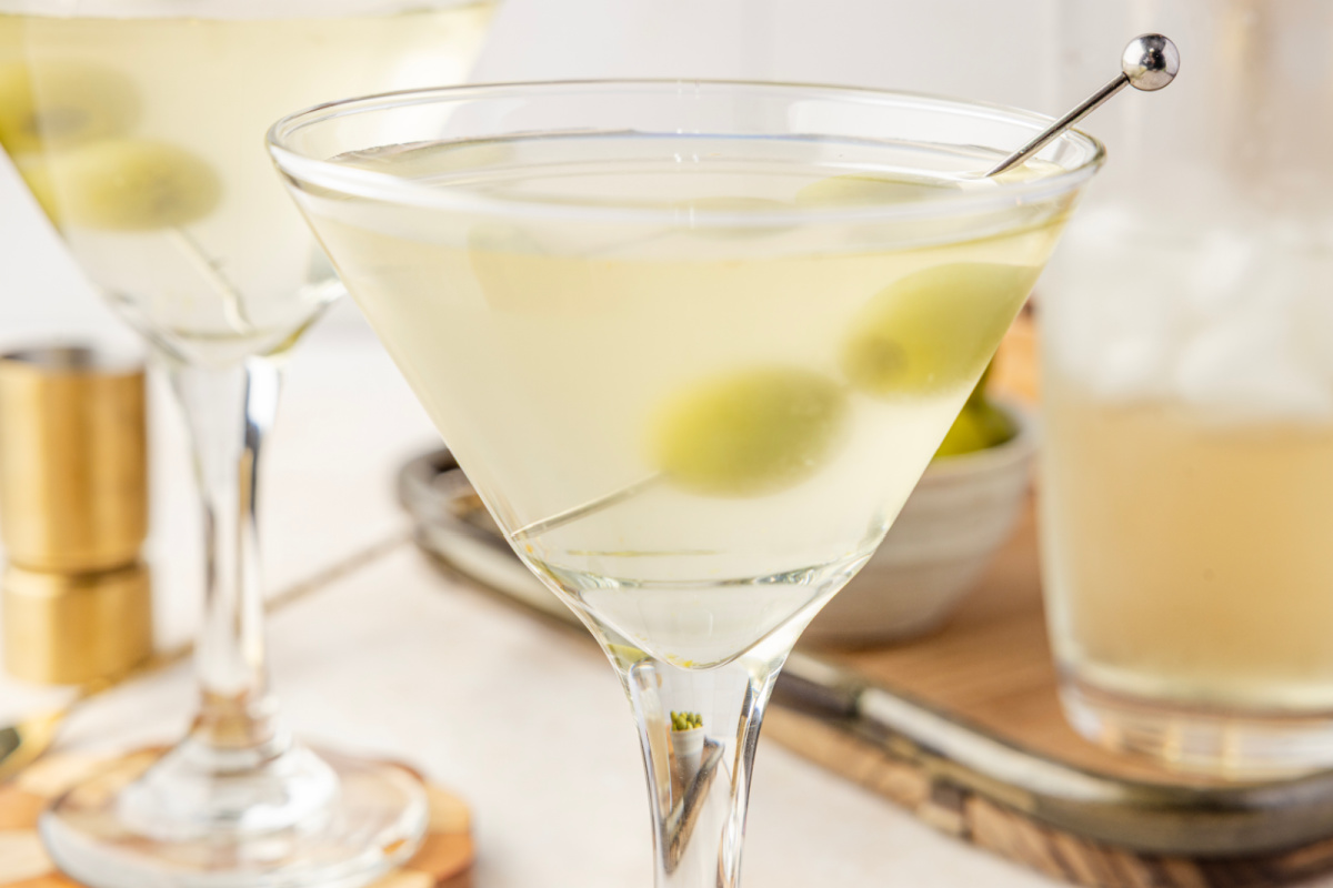 Two chilled martinis with green olives resting in pale, briny cocktails, set beside a shaker and a small bowl of olives.