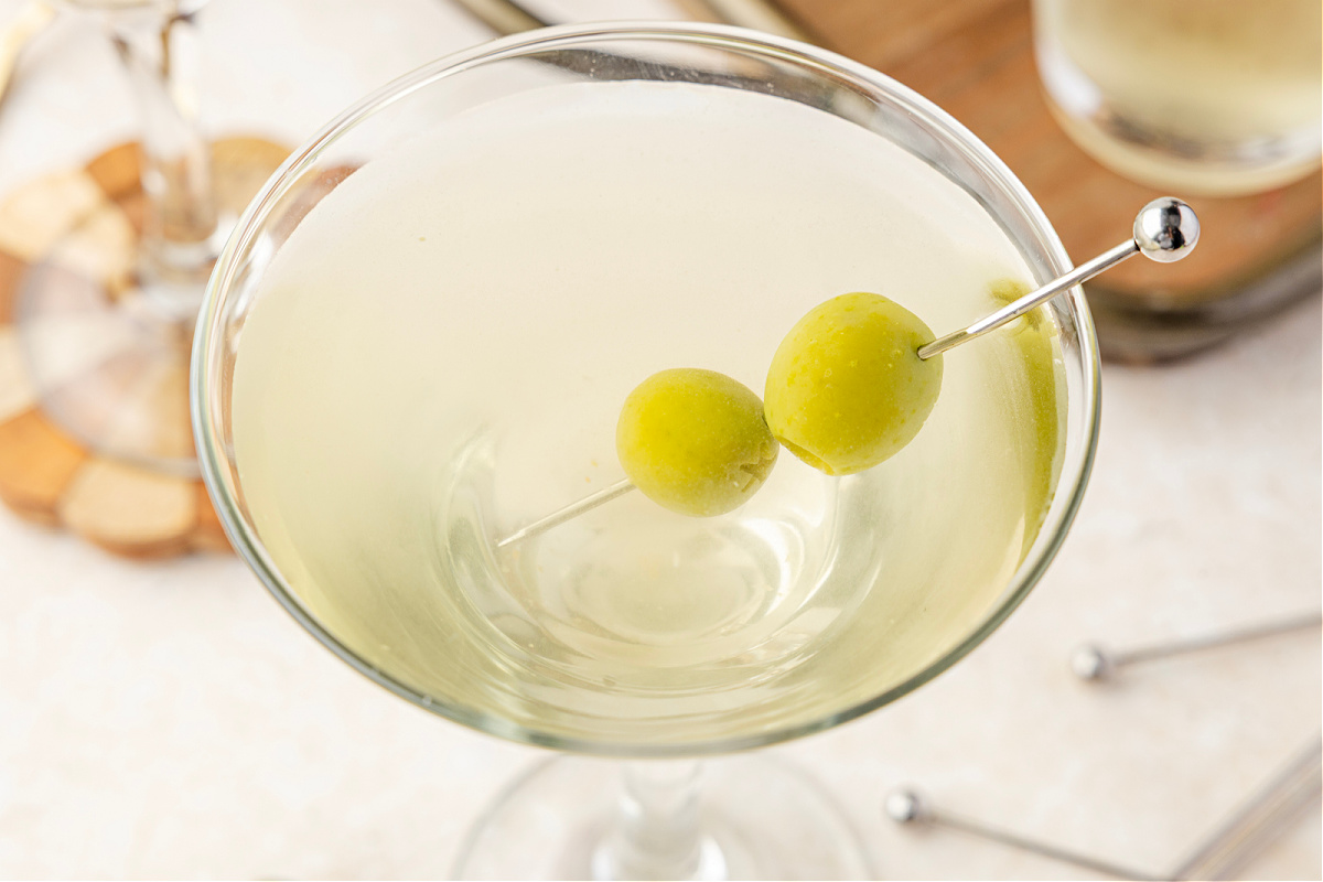 A chilled martini glass filled with a pale, briny cocktail and green olives, set against a shaker and bowls of olives in the background.