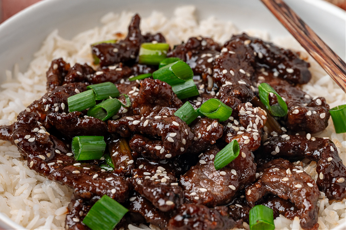 Close up of glossy sliced beef in a ginger soy sauce spooned over rice and finished with sesame seeds.