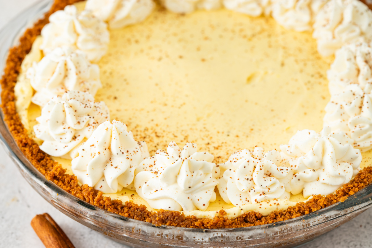A close-up of an eggnog cream pie with piped whipped cream around the edge, lightly dusted with nutmeg and set in a golden cookie crust.