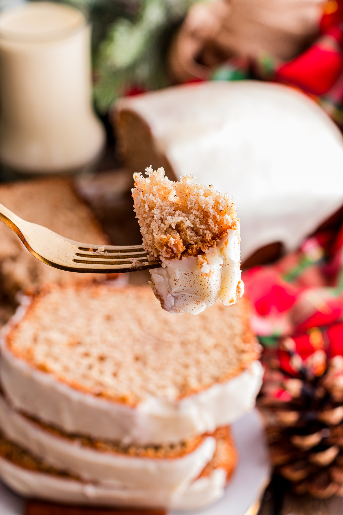 Fork holding a bite of eggnog bread with a soft, spiced crumb and set eggnog glaze, with the loaf blurred in the background on a holiday table.