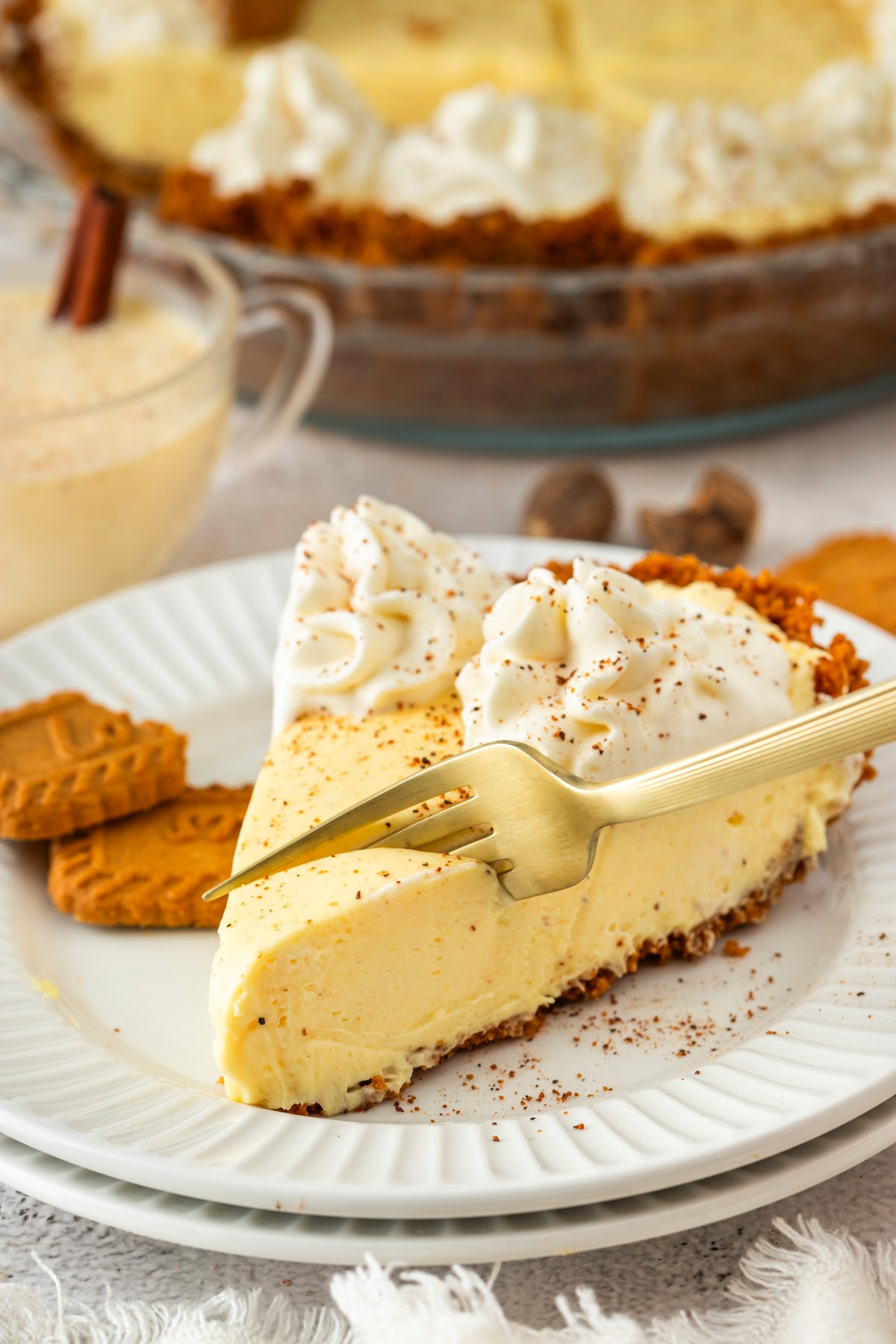 A plated slice of eggnog cream pie topped with whipped cream and nutmeg, served with a fork and cookies on the side.