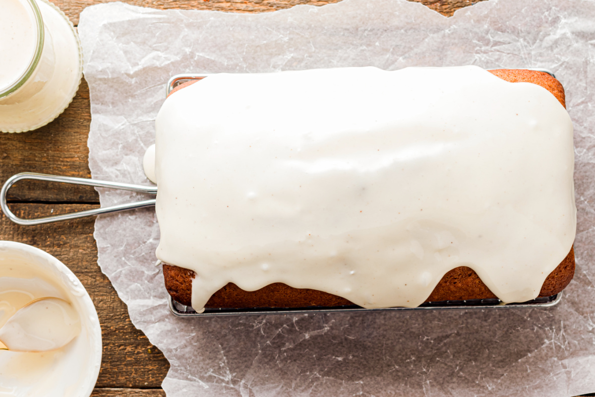 Freshly glazed eggnog quick bread resting on a cooling rack, with a smooth white glaze cascading over the top on parchment paper.