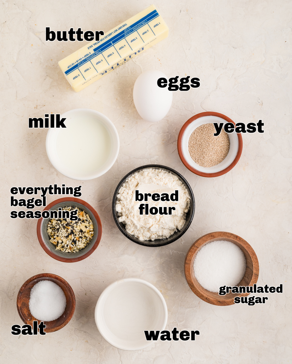 Measured ingredients for savory yeast rolls arranged on a light surface, including bread flour, yeast, eggs, milk, butter, sugar, salt, and everything bagel seasoning.