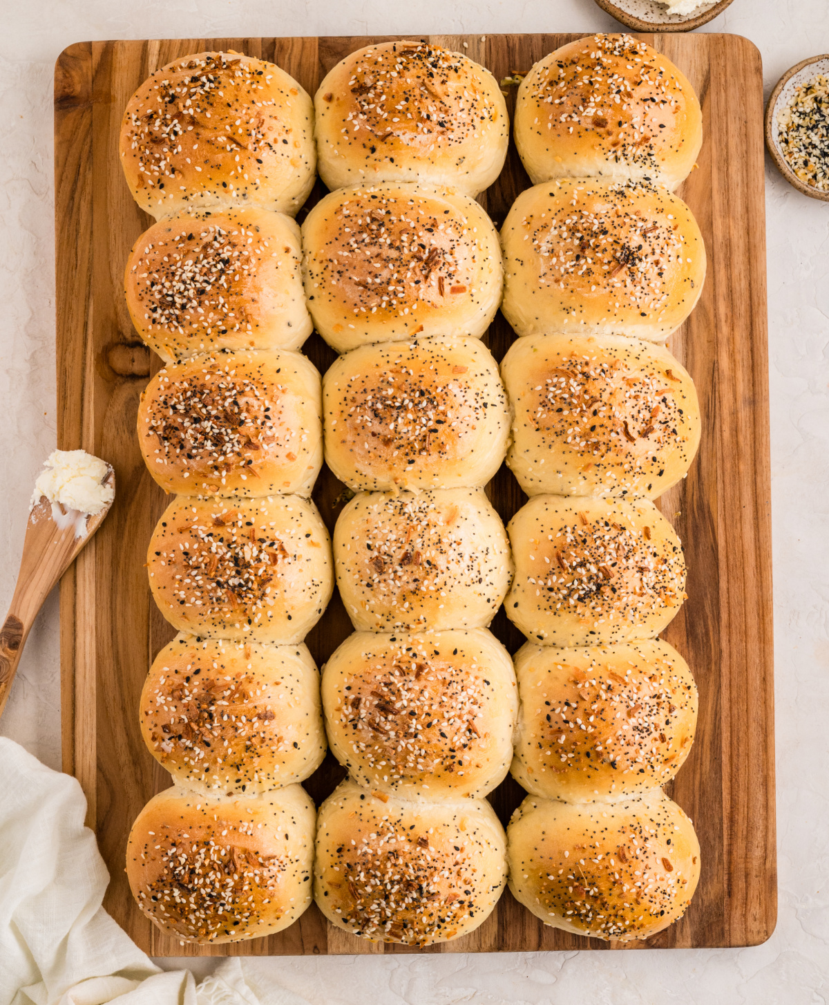 A full tray of golden baked rolls arranged in neat rows on a wooden board, topped with sesame seeds, poppy seeds, garlic, and onion seasoning.