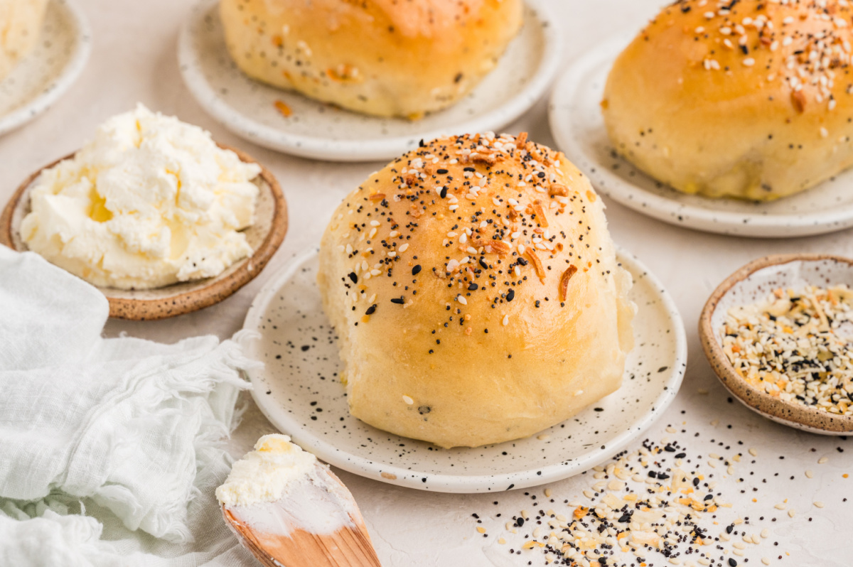 Soft baked rolls topped with sesame seeds, poppy seeds, garlic, and onion, served on small plates with cream cheese and extra seasoning nearby.