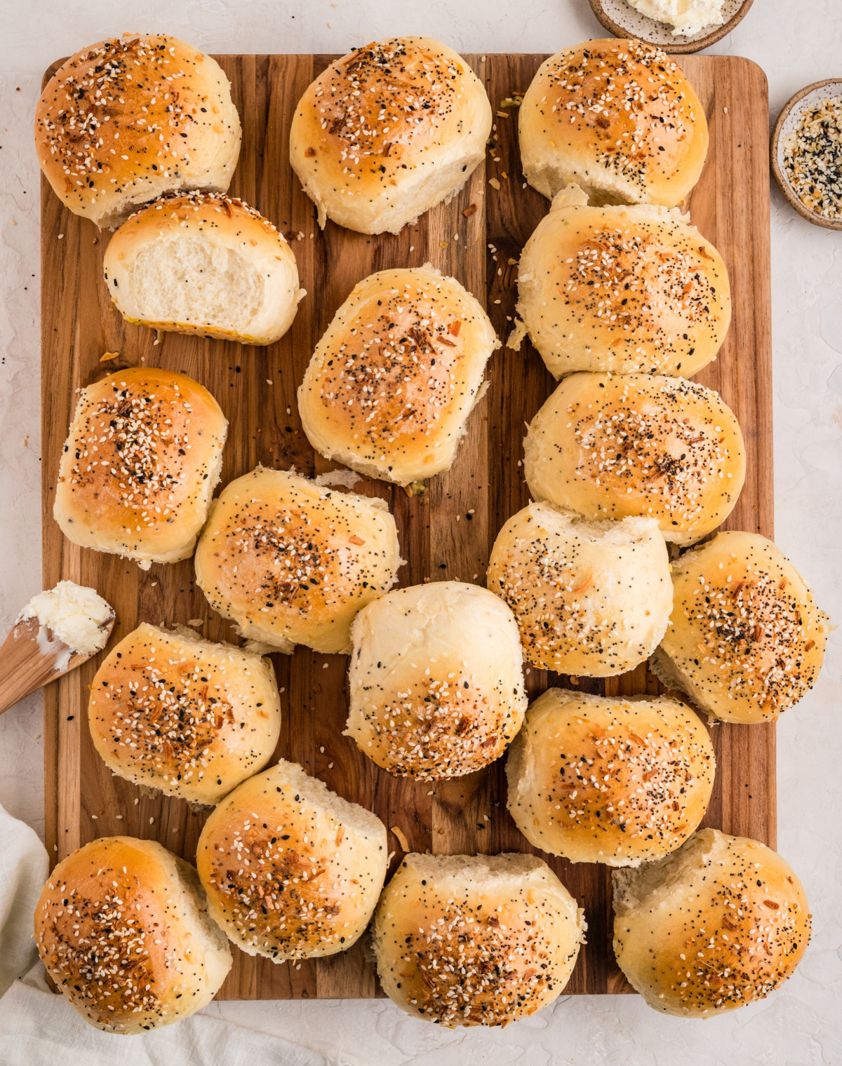 Golden baked rolls scattered across a wooden board, some split open to show the soft interior, with everything seasoning sprinkled over the tops.