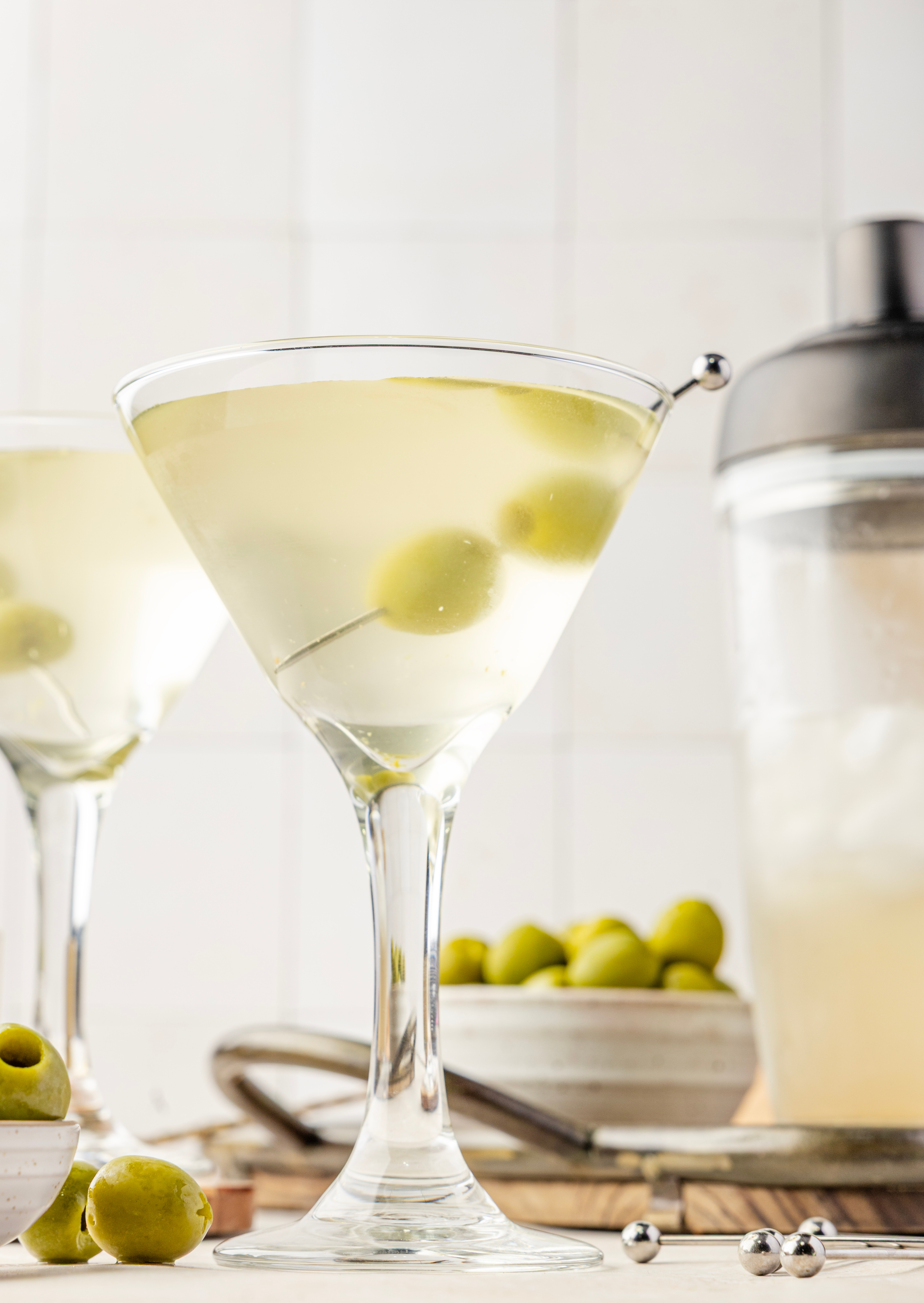 An extra dirty martini resting on a wooden tray, garnished with two green olives and surrounded by olive brine, ice, and cocktail picks.