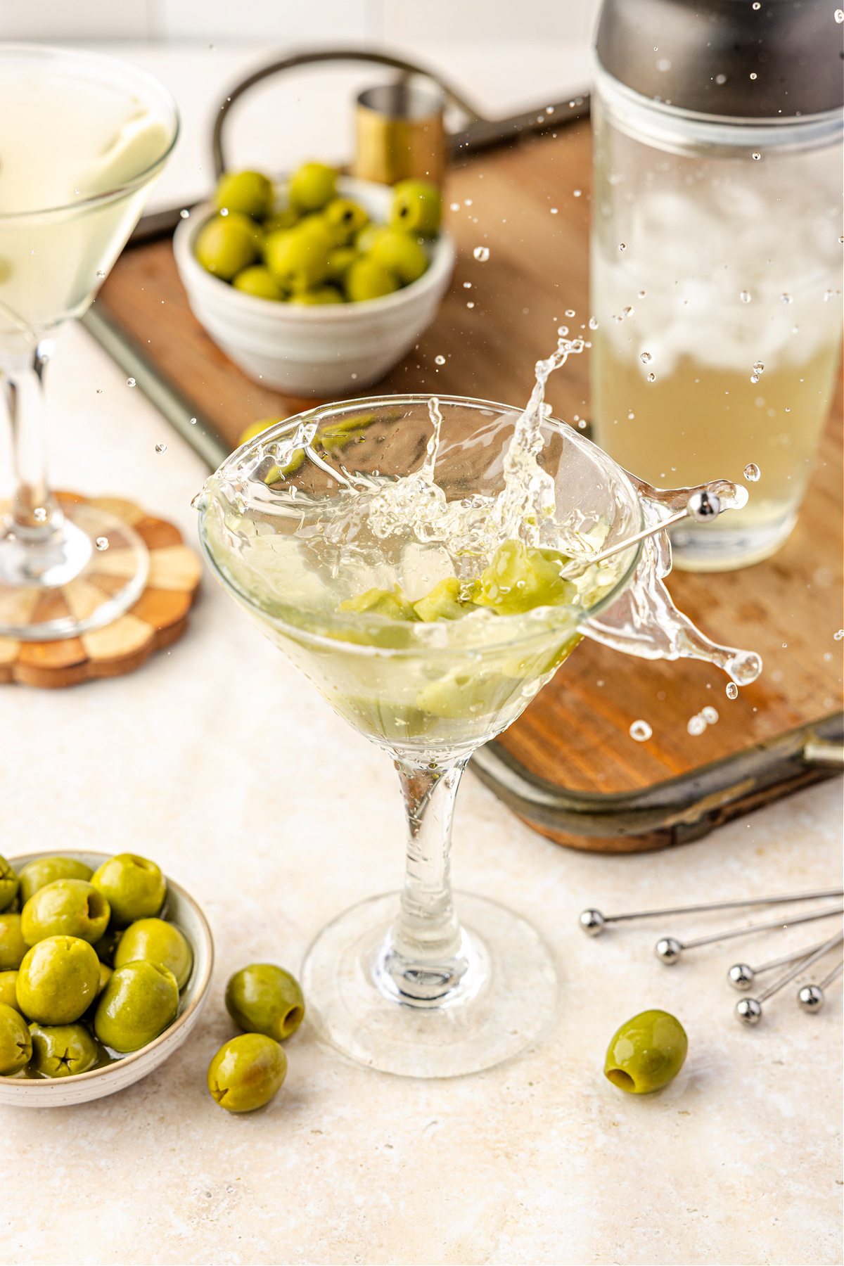 A chilled martini splashing dramatically in the glass as green olives tumble through the pale, briny cocktail on a bar tray.