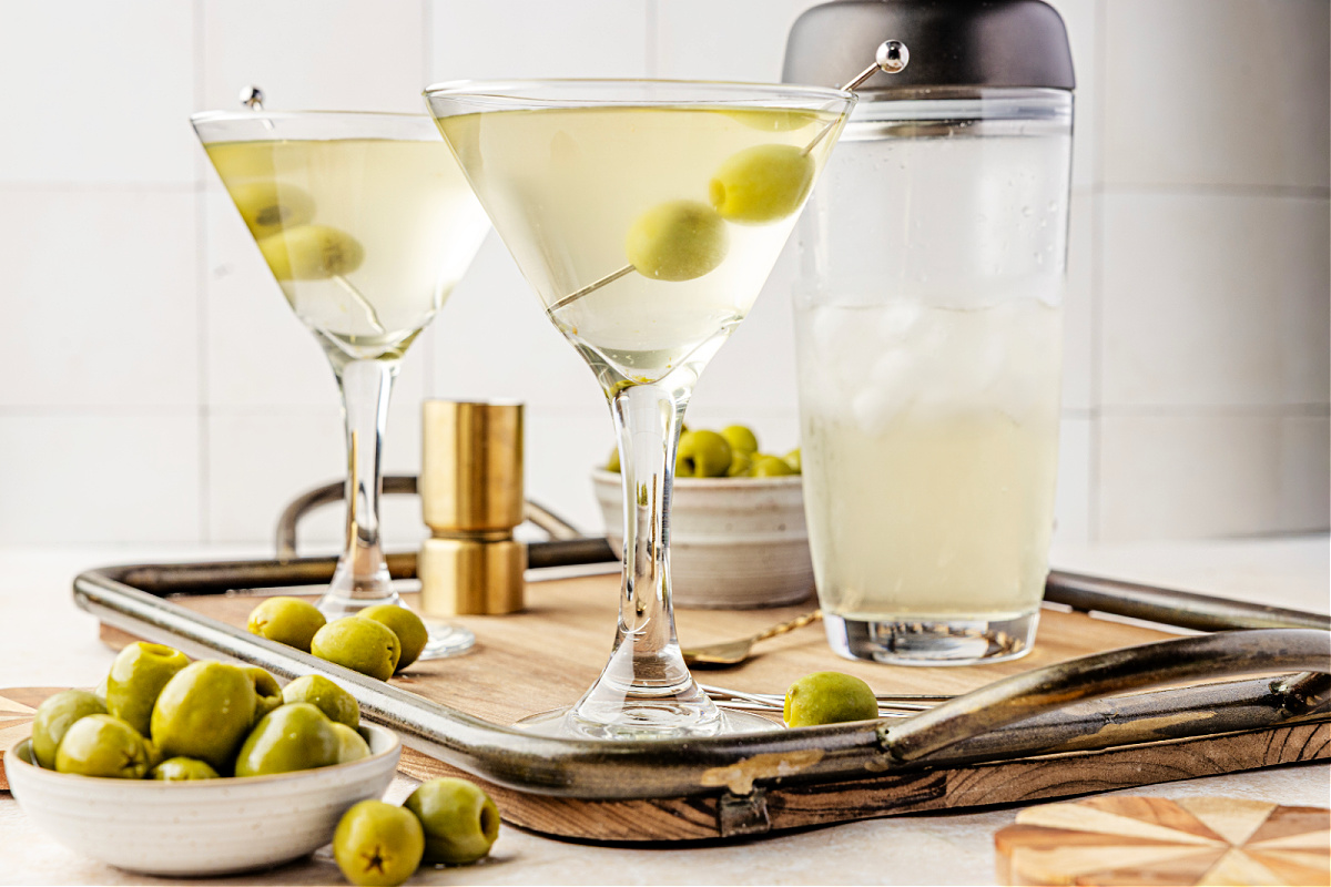 Two briny martinis garnished with green olives on a tray beside a cocktail shaker and bowls of olives.