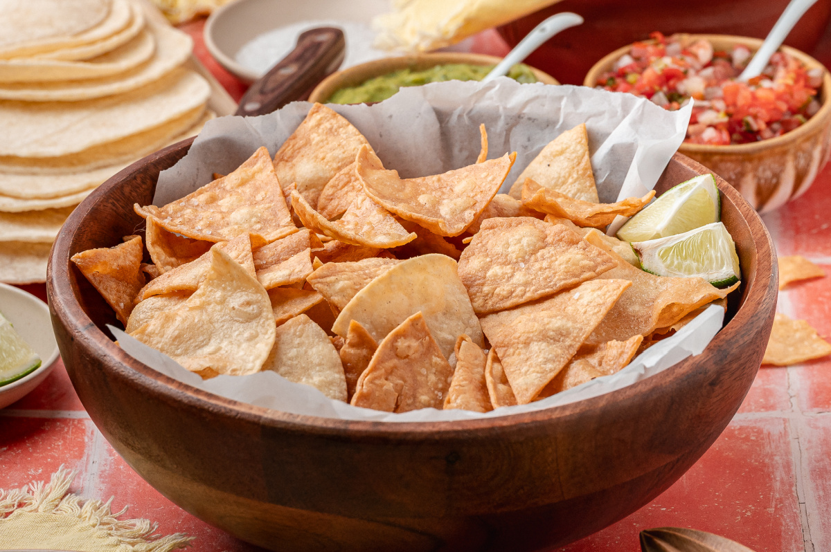 A wooden bowl filled with easy fried tortilla chips served with lime wedges and salsa nearby.