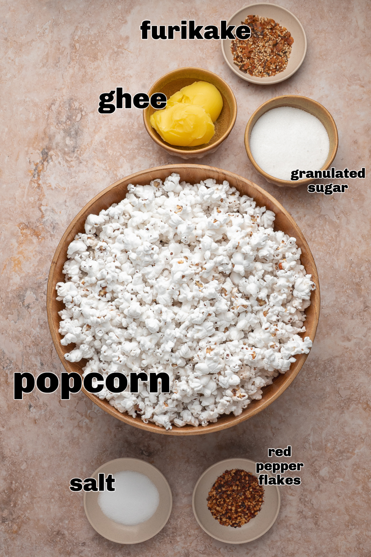 A flat lay of the popcorn ingredients arranged around a large wooden bowl, including ghee, sugar, salt, red pepper flakes, and furikake seasoning.
