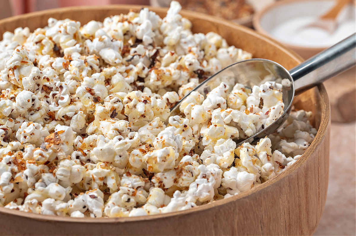 A close-up of seasoned popcorn in a wooden bowl, with a metal scoop resting against the side.