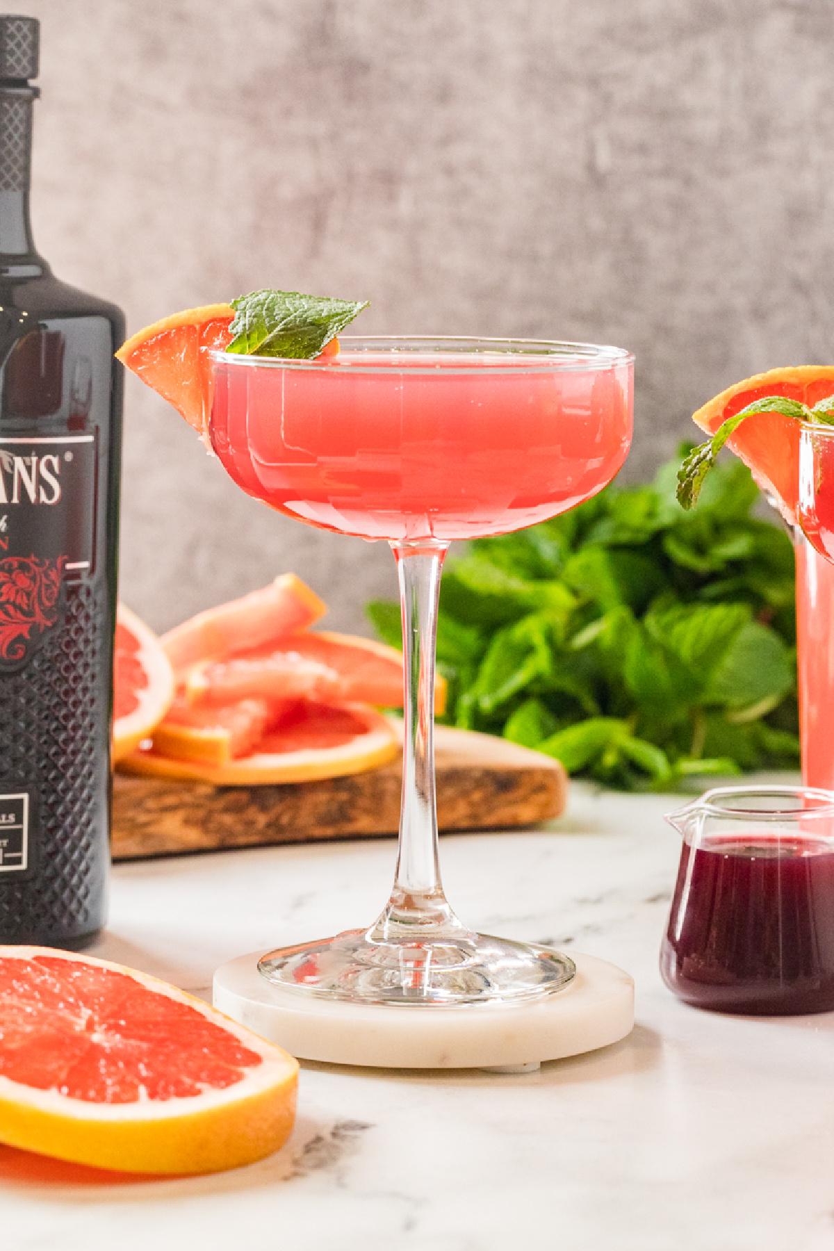 A pink grapefruit and pomegranate cocktail served in a coupe glass, garnished with a grapefruit slice and mint, with fresh citrus and syrup in the background.