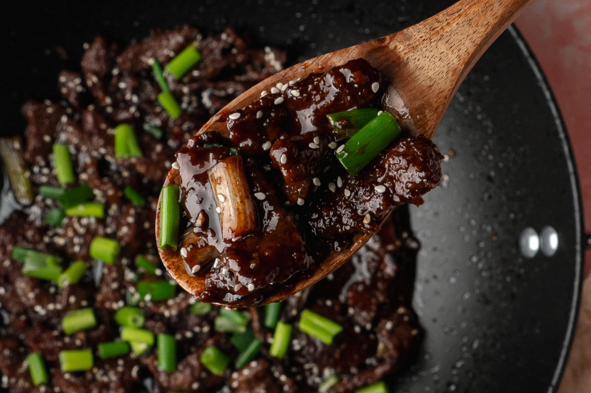 A wooden spoon lifting saucy slices of beef with green onions and sesame seeds from a hot skillet.