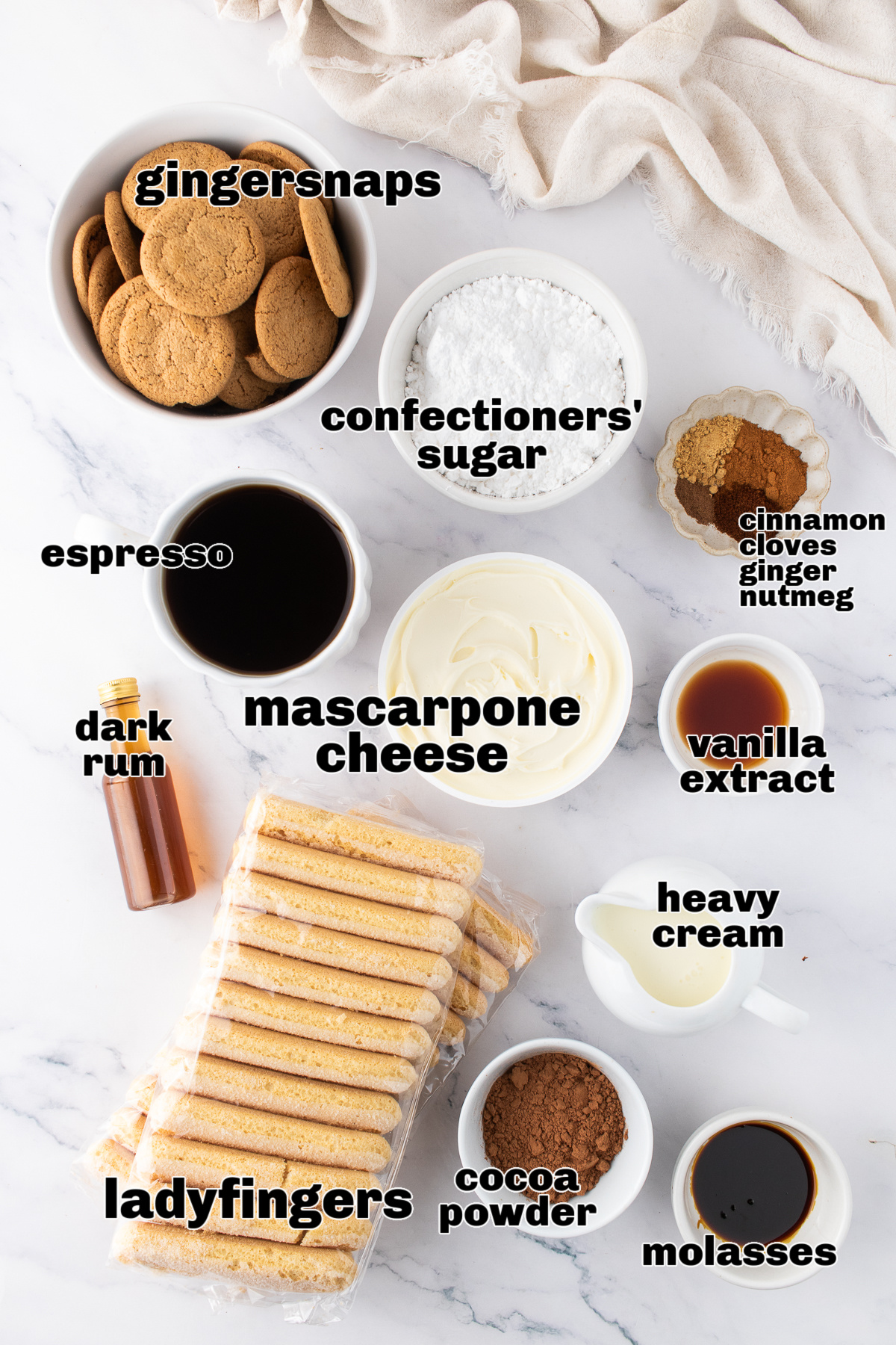 A top-down view of the gingerbread tiramisu ingredients laid out on a marble surface, including gingersnaps, ladyfingers, mascarpone, espresso, spices, molasses, and confectioners&rsquo; sugar.
