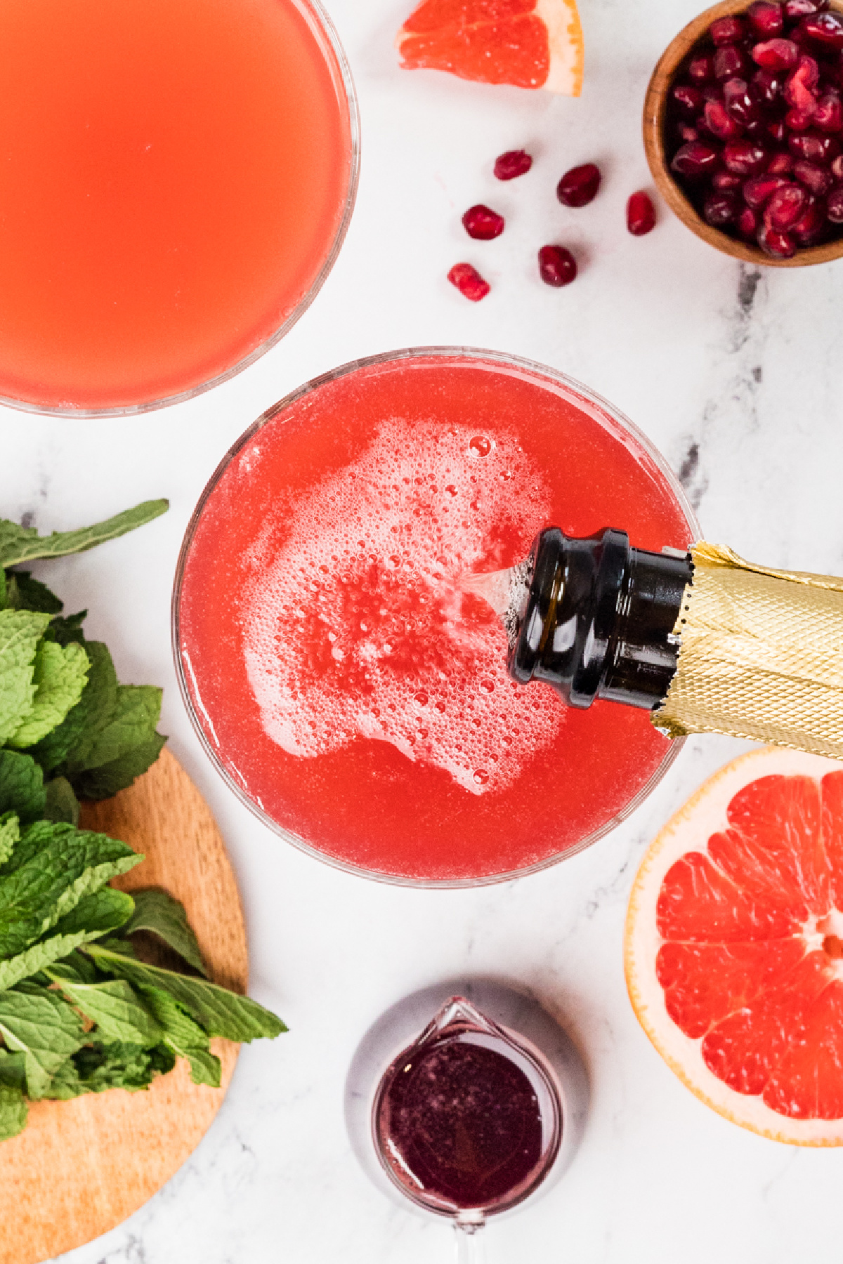 Top-down view of a grapefruit pomegranate gin fizz being topped with prosecco, showing fine bubbles forming over the pink cocktail with fresh grapefruit, pomegranate arils, and mint nearby.
