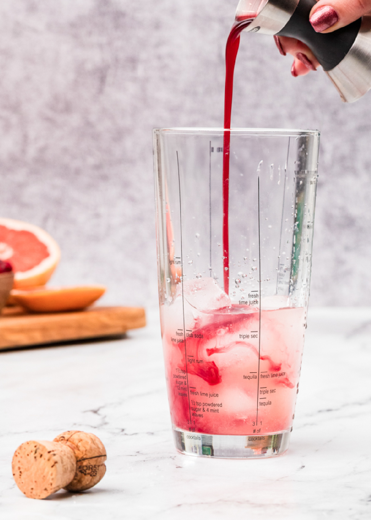 Pomegranate simple syrup being poured into a cocktail shaker filled with ice and grapefruit juice, showing the deep red color as it blends.
