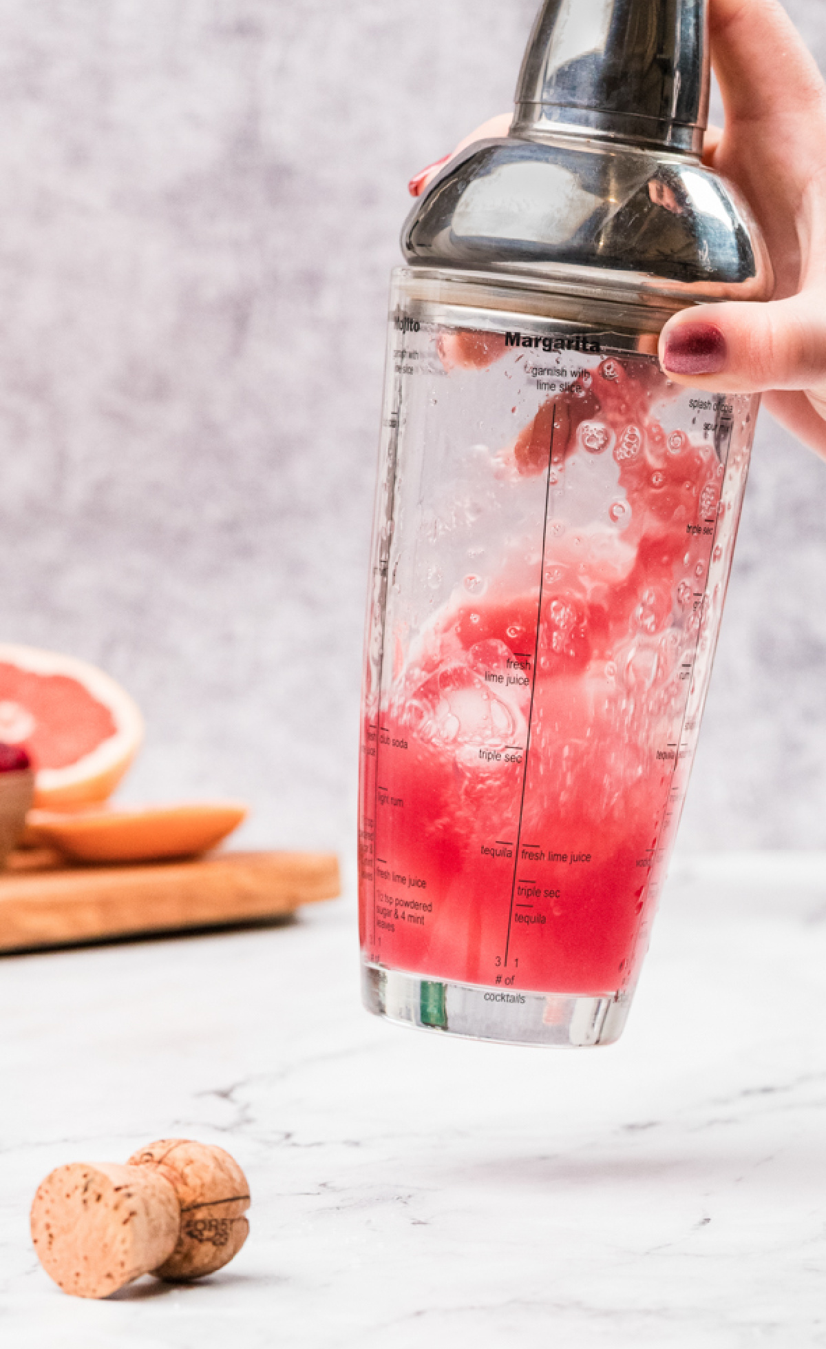 A cocktail shaker being shaken with ice, grapefruit juice, gin, and pomegranate syrup, showing the pink mixture swirling inside.