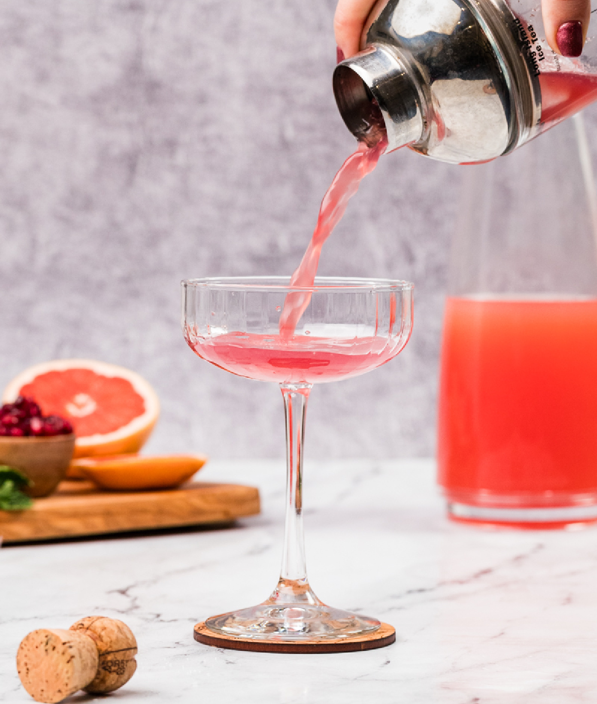 Straining a pink grapefruit and pomegranate cocktail into a coupe glass, showing the clear pour and pale rosy color before adding prosecco.
