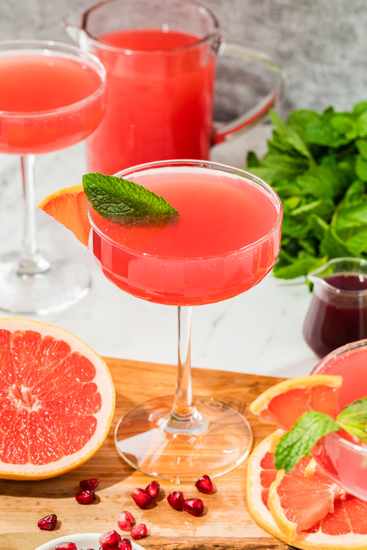 Coupe glasses filled with a pink grapefruit and pomegranate cocktail on a wooden board, surrounded by fresh grapefruit halves, pomegranate arils, mint, and a pitcher of juice.