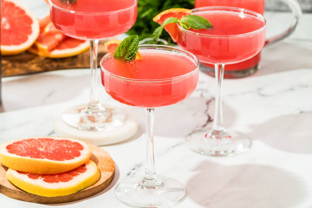Coupe glasses filled with a pink grapefruit and pomegranate gin cocktail, garnished with fresh grapefruit slices and mint on a marble surface.