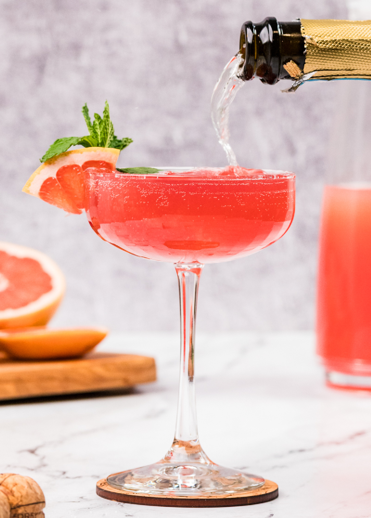 A grapefruit pomegranate gin fizz being topped with prosecco in a coupe glass, showing the deep pink color, fine bubbles, and a garnish of grapefruit and mint.