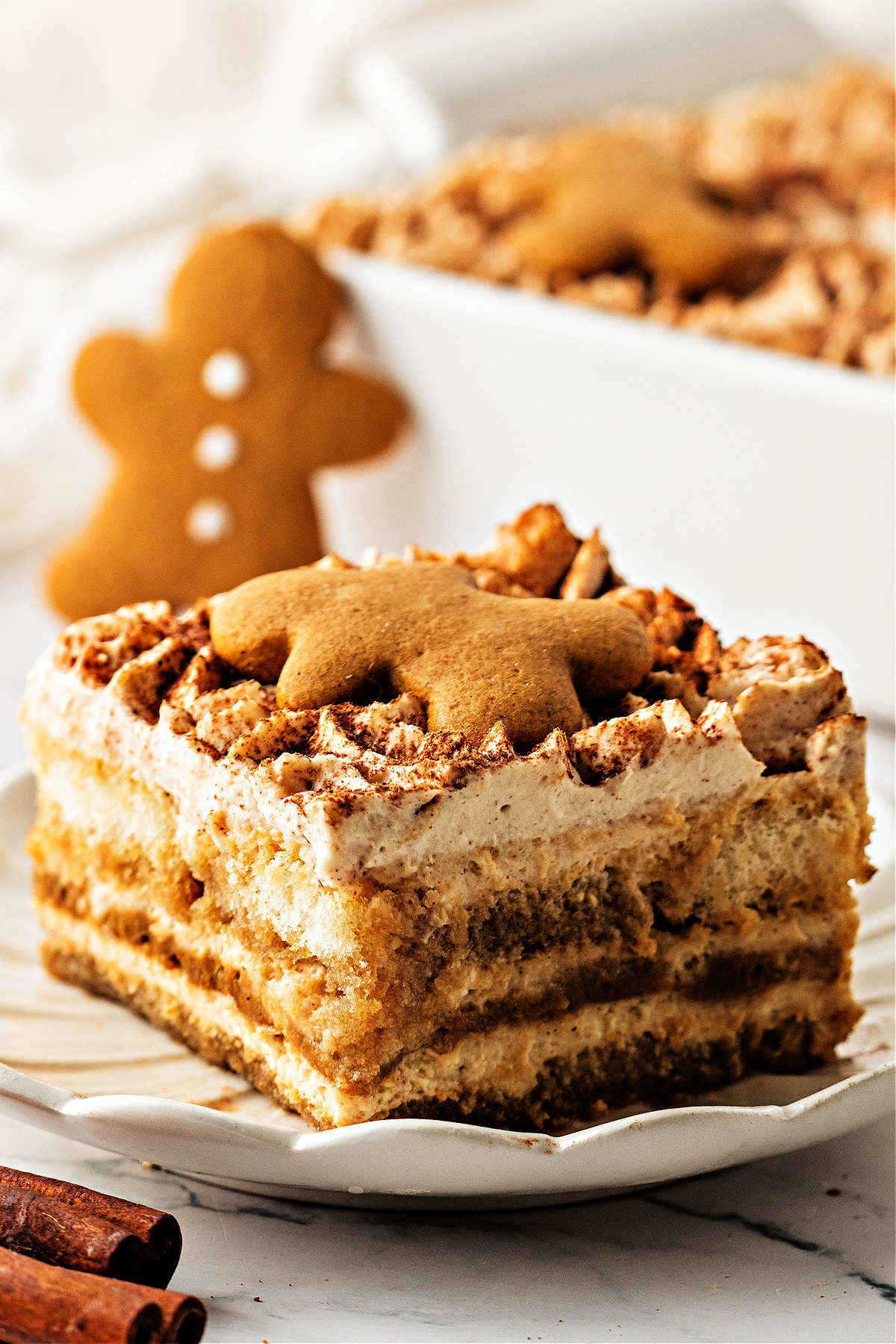 A close slice of gingerbread tiramisu on a scalloped plate, layered with espresso-soaked ladyfingers and spiced mascarpone, topped with a small gingerbread cookie and dusted with cocoa.