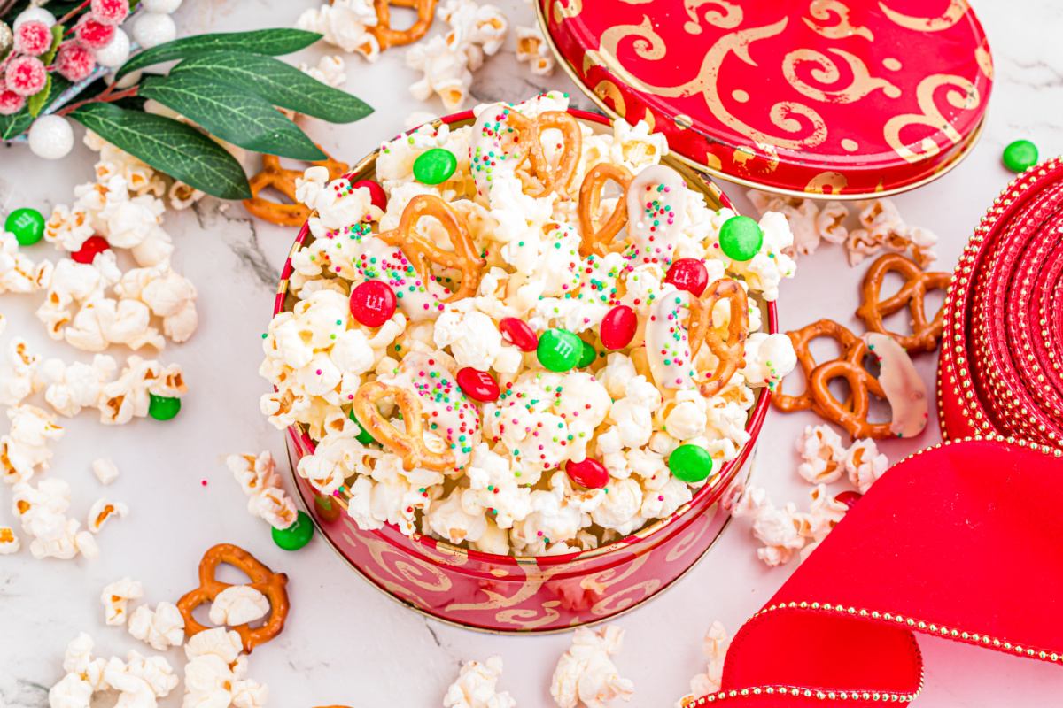 Festive popcorn mix with white chocolate&ndash;dipped pretzels, red and green candies, and sprinkles in a holiday tin.