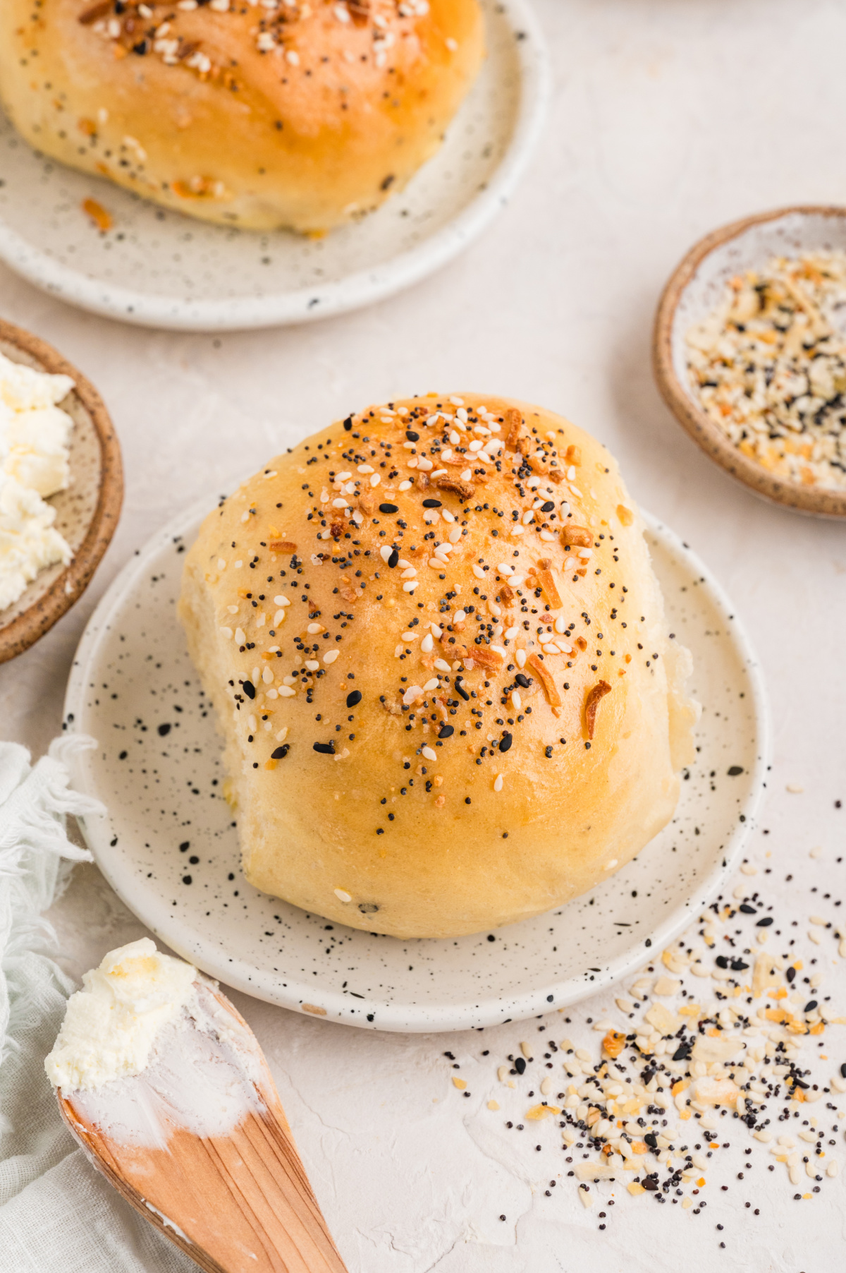 A single golden baked roll topped with sesame seeds, poppy seeds, garlic, and onion, resting on a small plate with cream cheese and loose seasoning nearby.