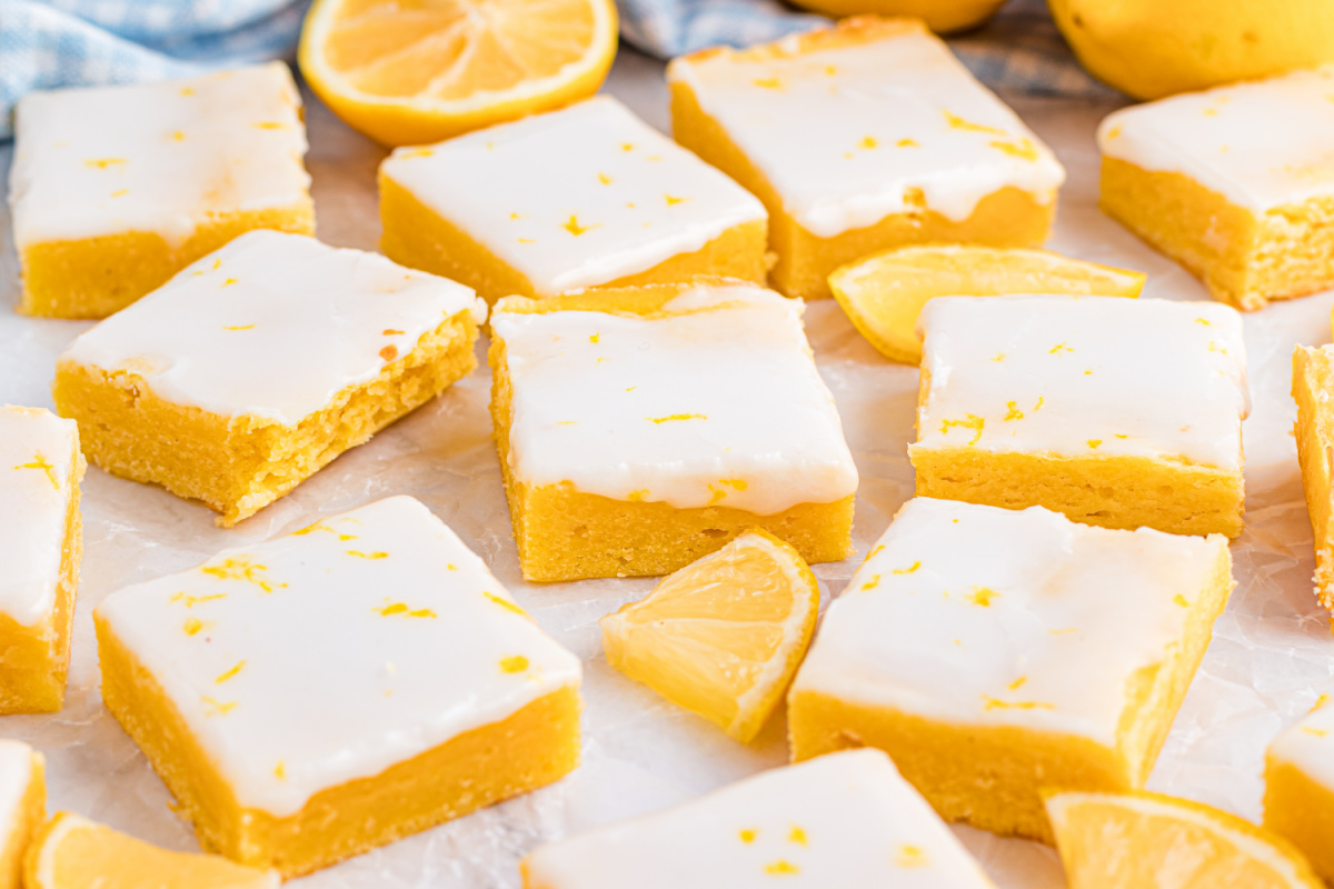 Lemon blondies arranged in a loose grid on parchment with fresh lemons nearby, highlighting the pale glaze and bright citrus color.