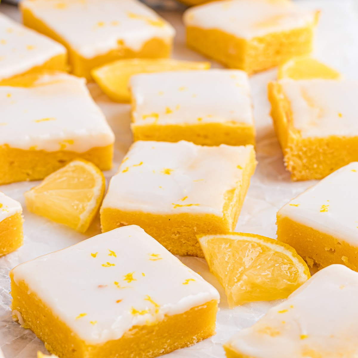 Lemon blondies arranged on parchment with a thin lemon glaze and fresh lemon wedges, showing clean edges and a dense, buttery interior.