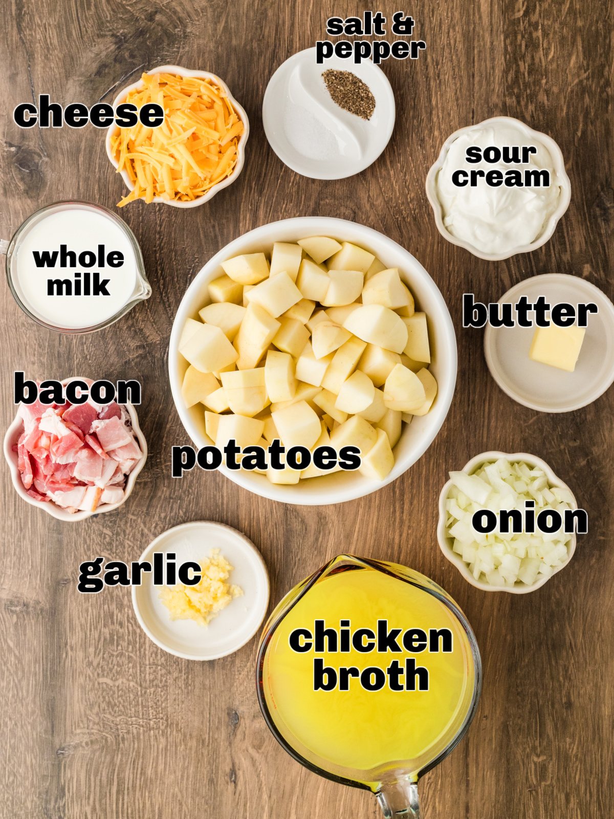 Ingredients for potato soup arranged on a wooden surface, including cubed potatoes, bacon, onion, garlic, cheese, sour cream, milk, butter, and chicken broth.