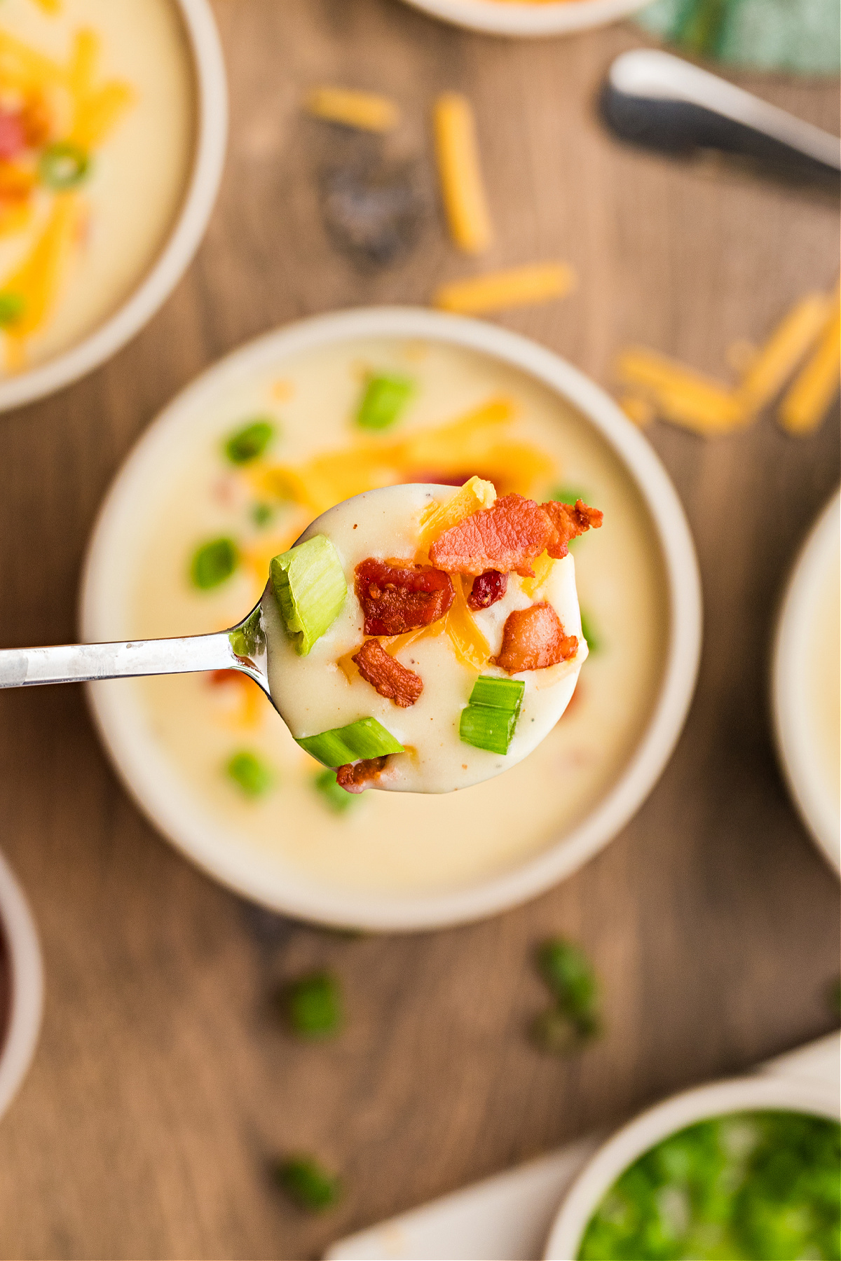 A spoonful of creamy potato soup topped with crisp bacon, melted cheddar, and chopped scallions held over a bowl.