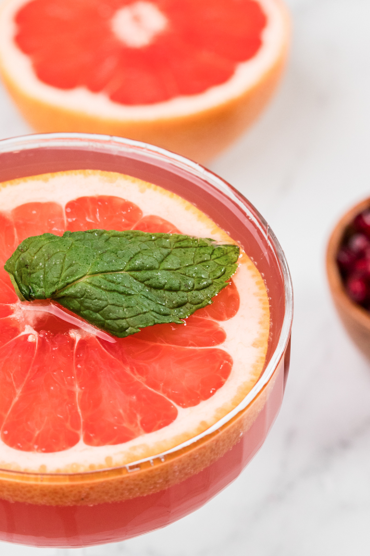 Close-up of a pink grapefruit and pomegranate cocktail garnished with a floating grapefruit slice and fresh mint, showing the clear rim and saturated winter color.