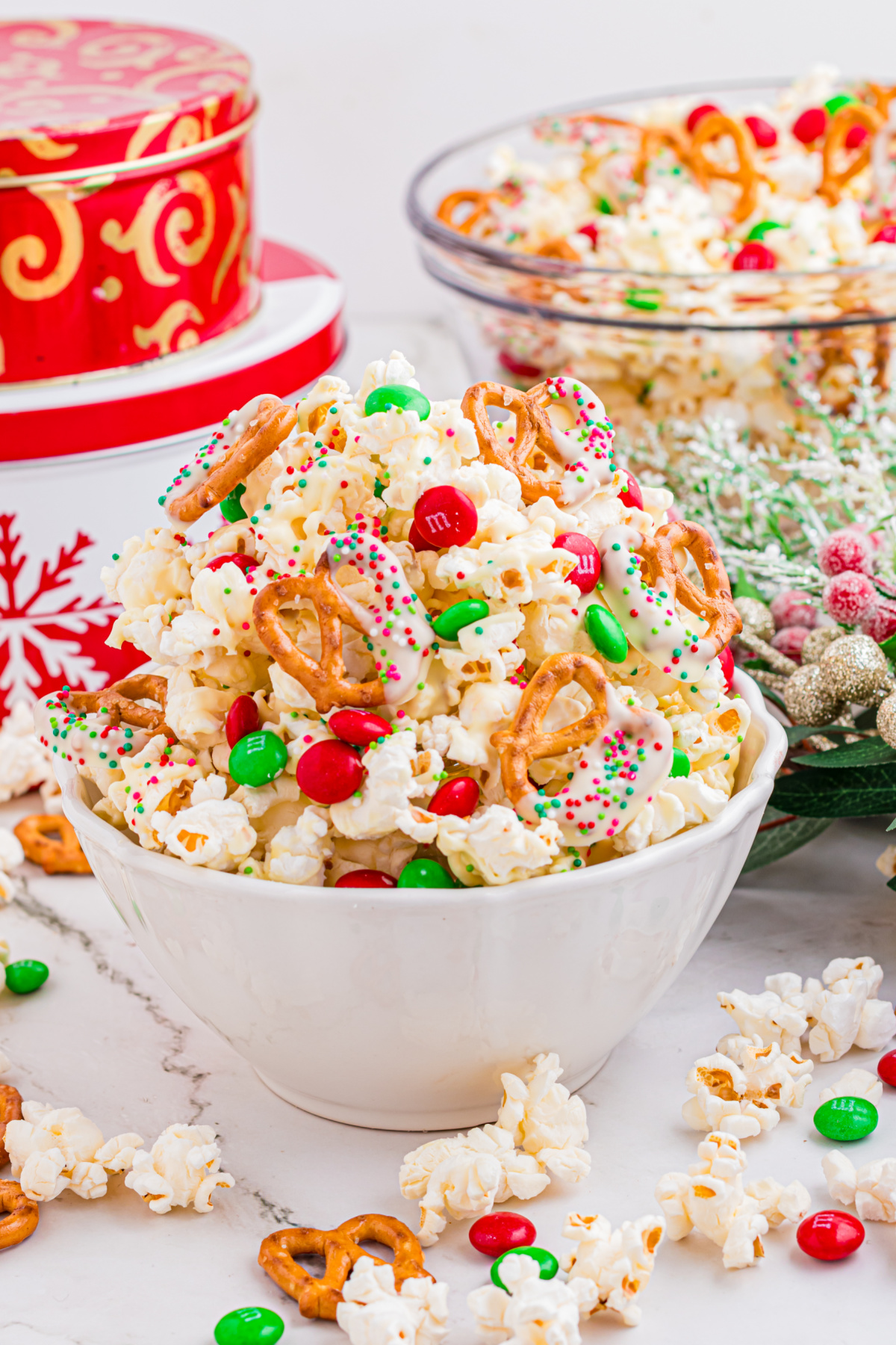 Christmas popcorn mix piled into a white bowl with white chocolate&ndash;dipped pretzels, red and green candies, and holiday sprinkles.