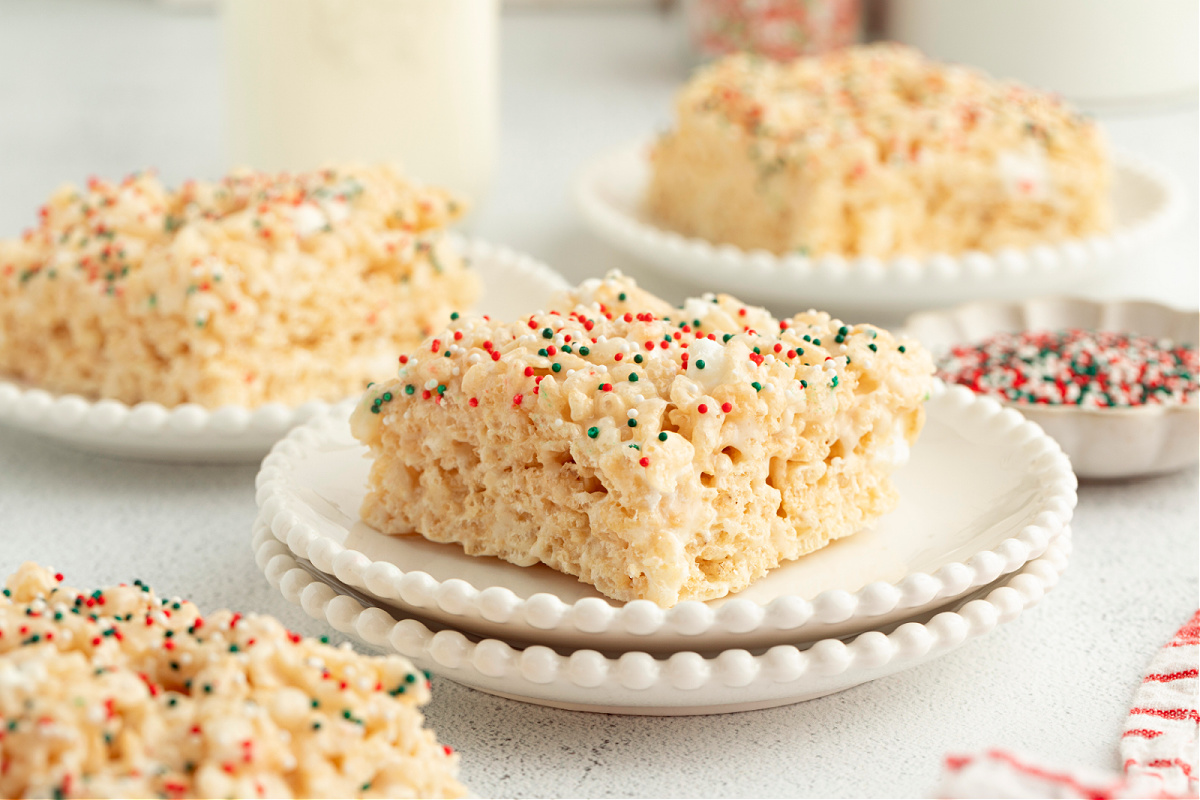 Soft sugar cookie&ndash;flavored Rice Krispie treat squares stacked on white plates with festive holiday sprinkles.