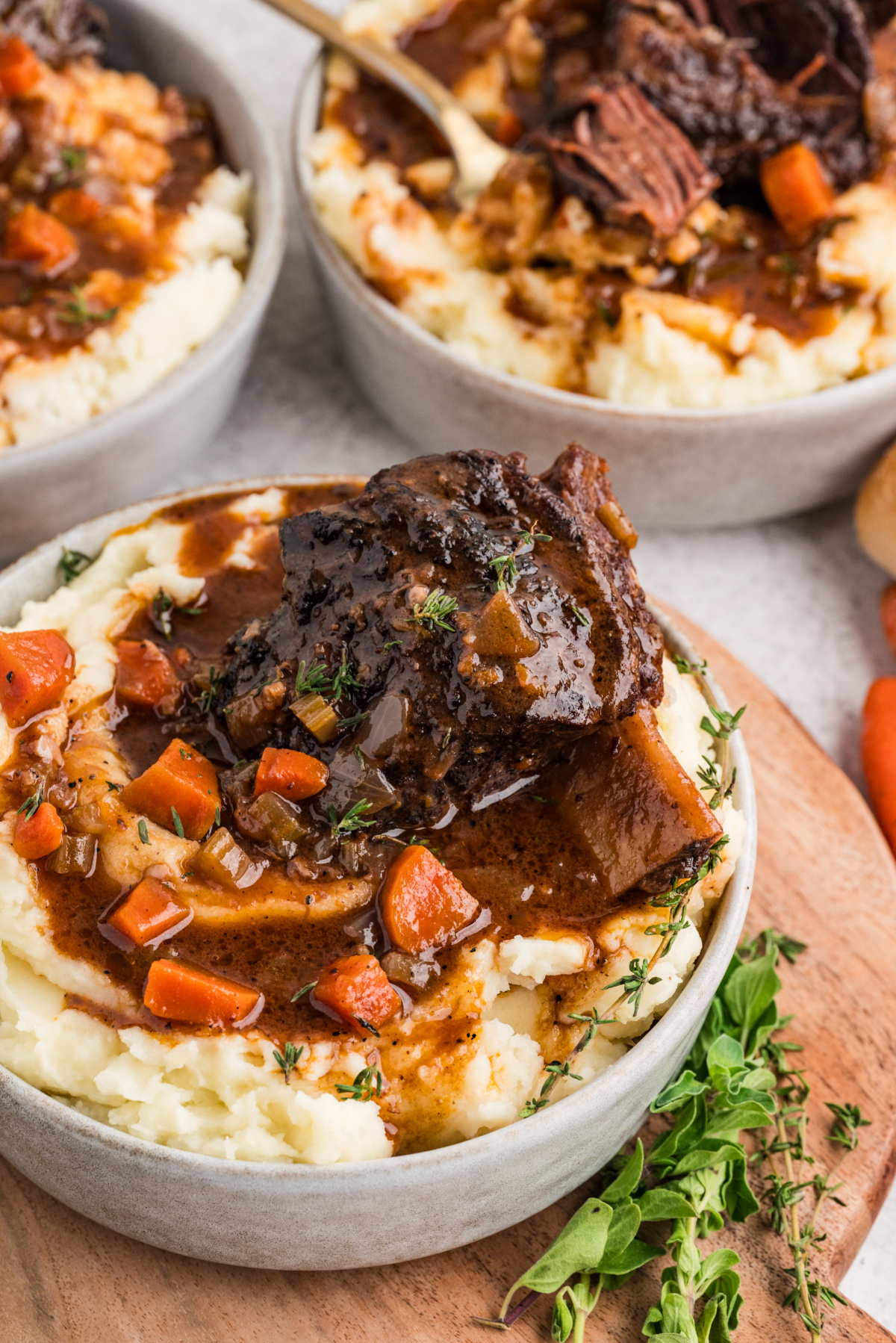 A bowl of oven-braised short ribs resting on mashed potatoes, spooned with glossy sauce and scattered with carrots and fresh herbs.