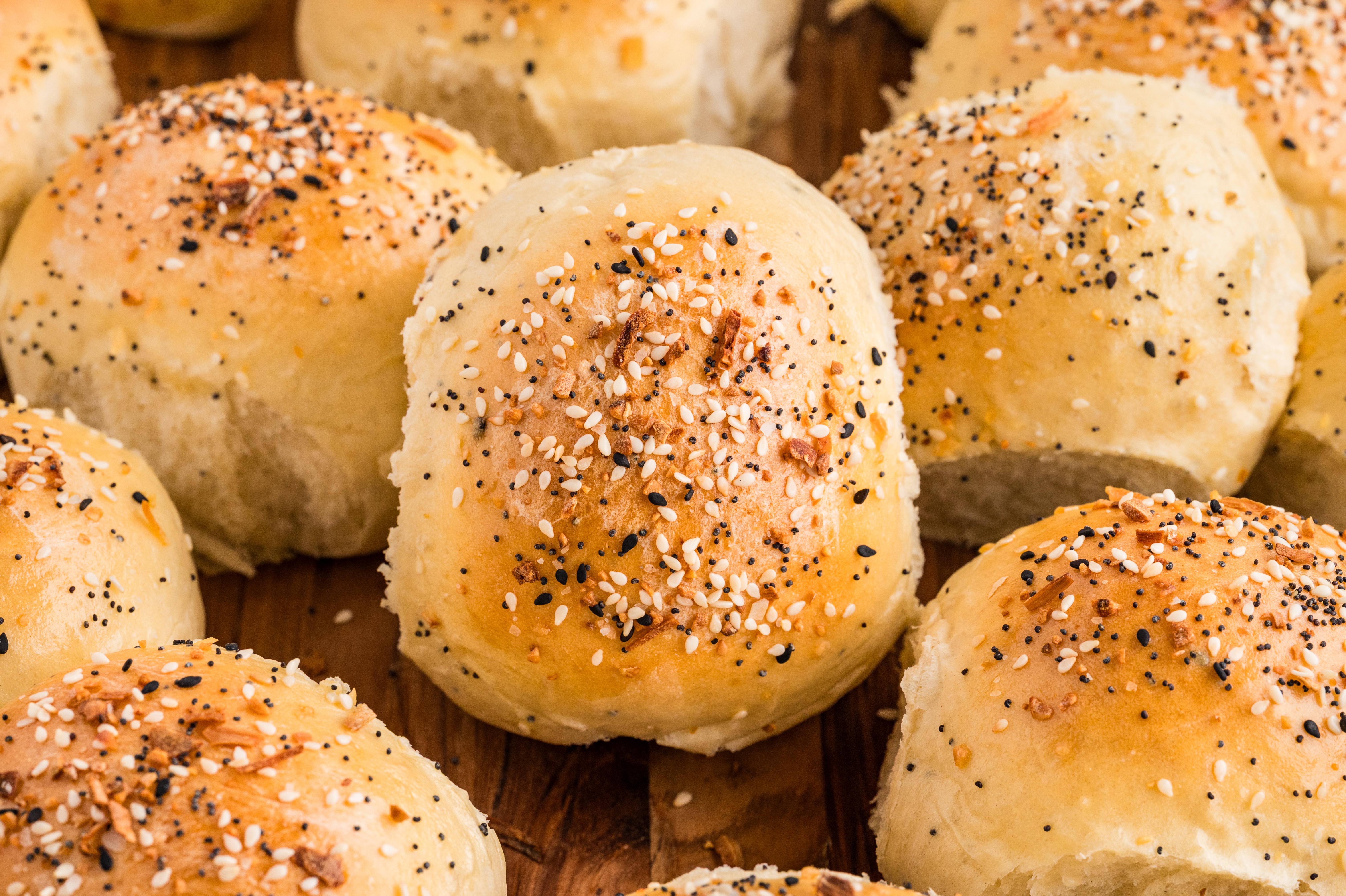 Golden baked rolls arranged on a wooden board, topped with sesame seeds, poppy seeds, garlic, and onion, with one roll split open to show the soft interior.