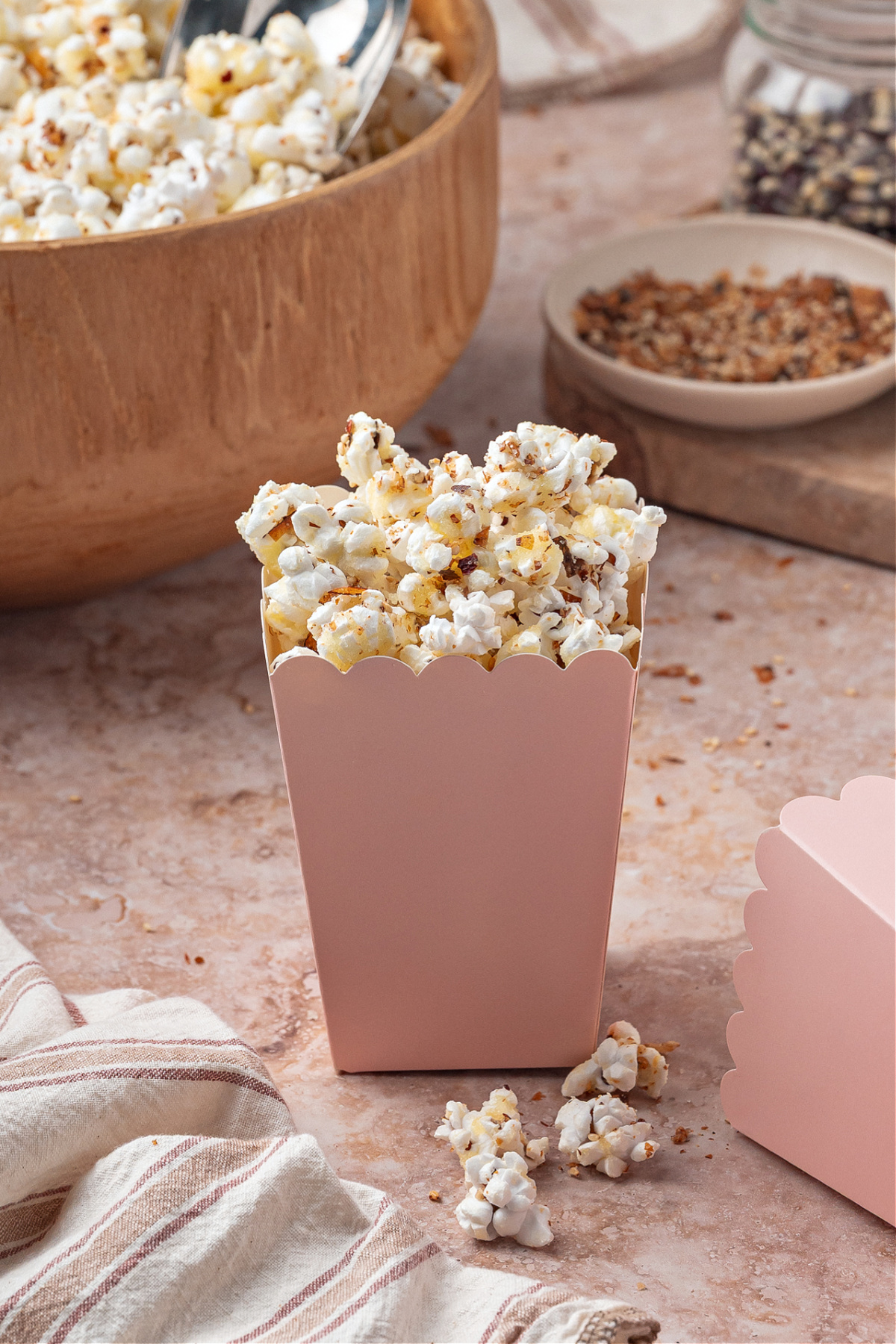 A pink snack box filled with sweet and spicy seasoned popcorn sits on a stone surface with a wooden bowl of popcorn in the background.