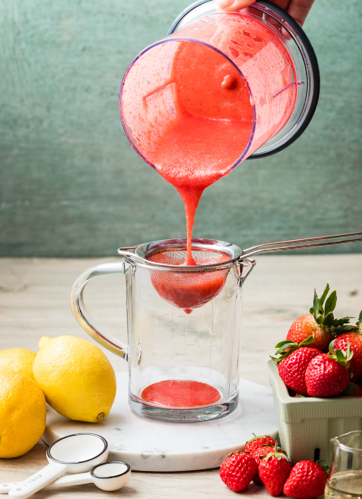 Fresh strawberry pur&eacute;e being strained through a fine-mesh sieve into a glass measuring cup.