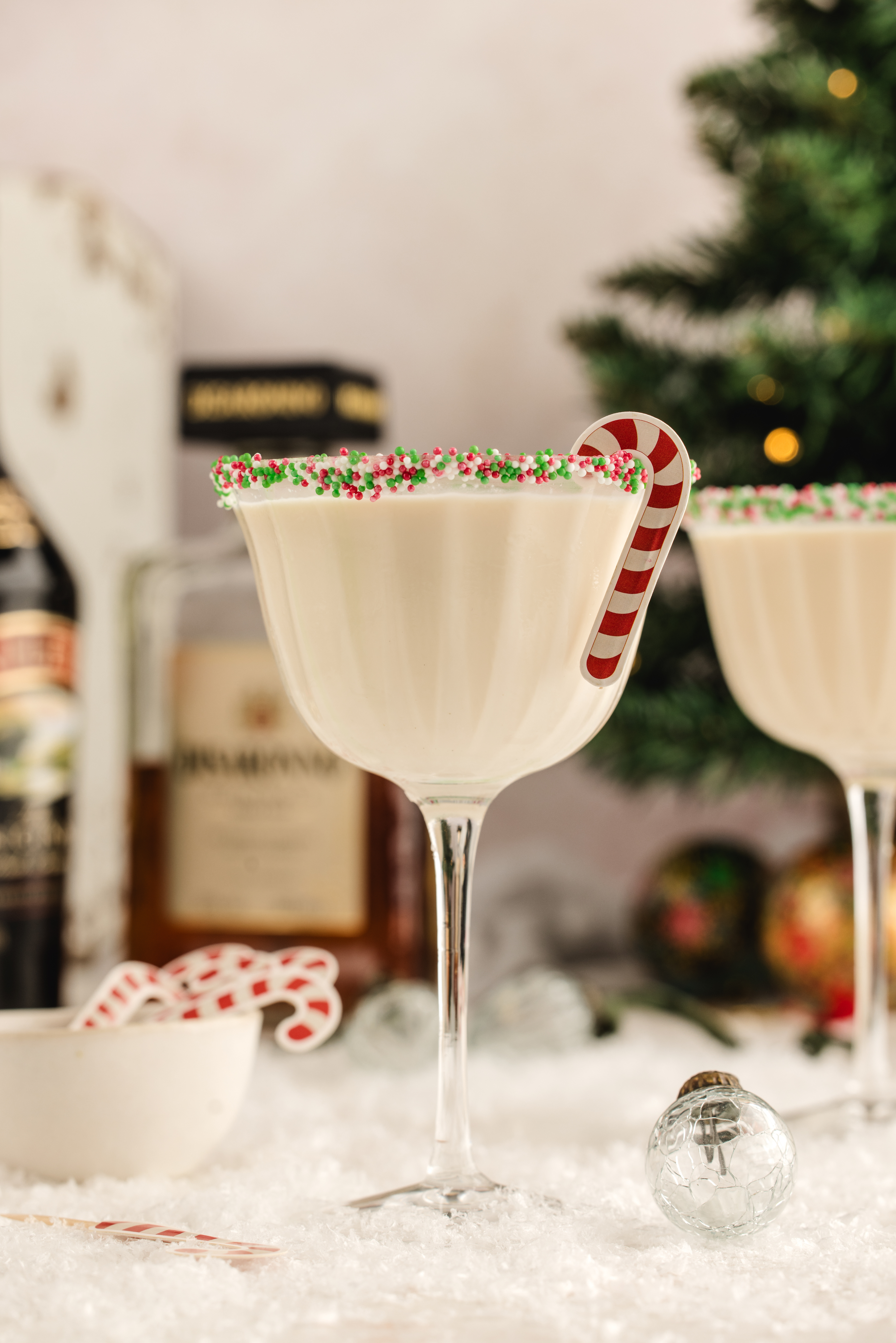 A creamy holiday cocktail in a sprinkle-rimmed coupe with a candy cane garnish, surrounded by snowy d&eacute;cor and bottles in the background.