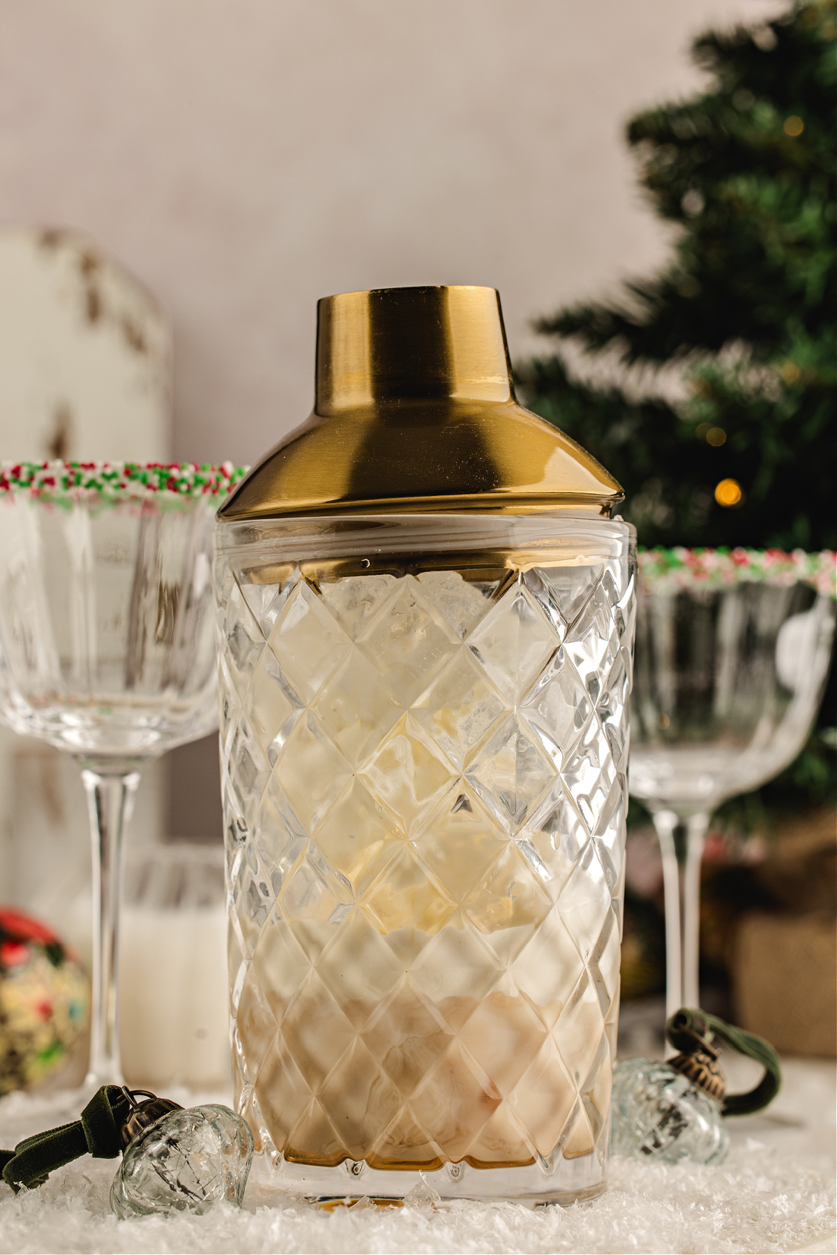 Irish cream being poured from a gold jigger into a mixing glass filled with ice and amaretto, with sprinkle-rimmed holiday coupes in the background.