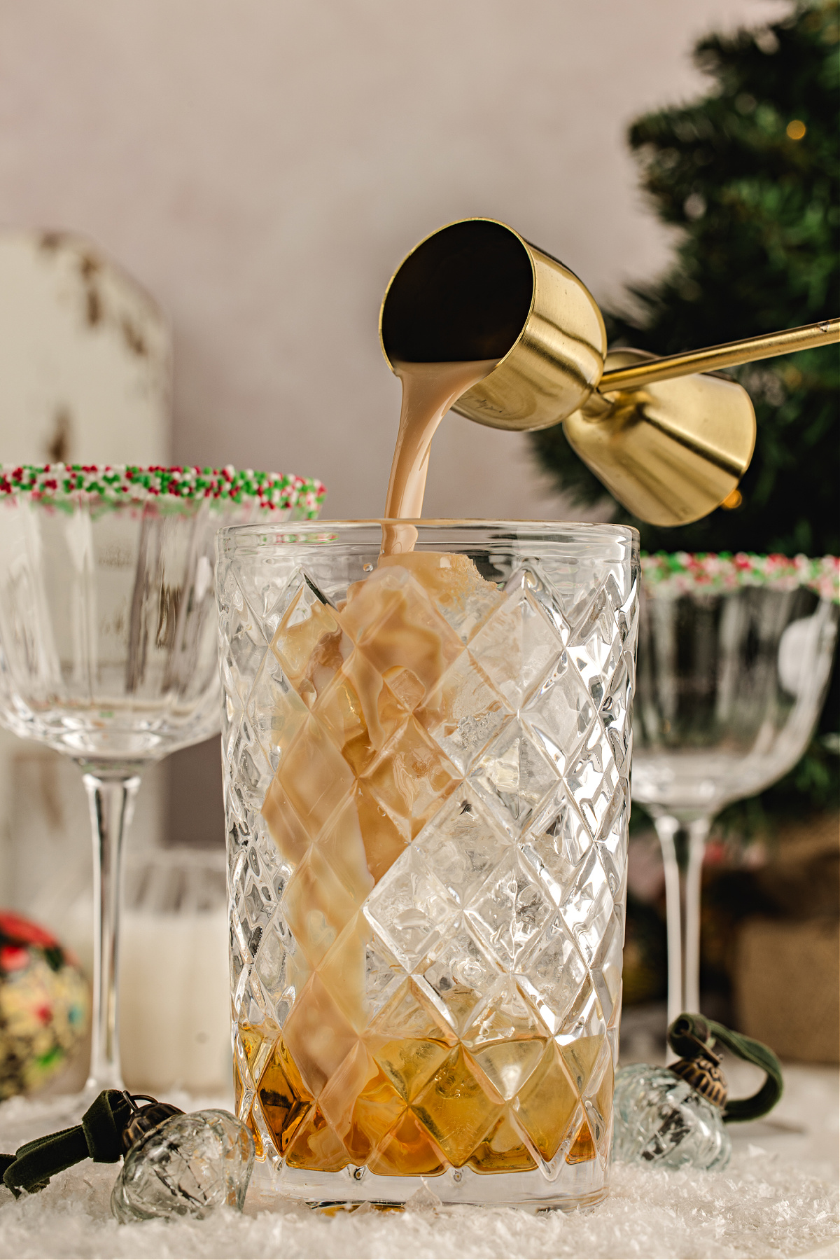 Irish cream being poured from a gold jigger into a mixing glass filled with ice and amaretto, with sprinkle-rimmed holiday coupes in the background.