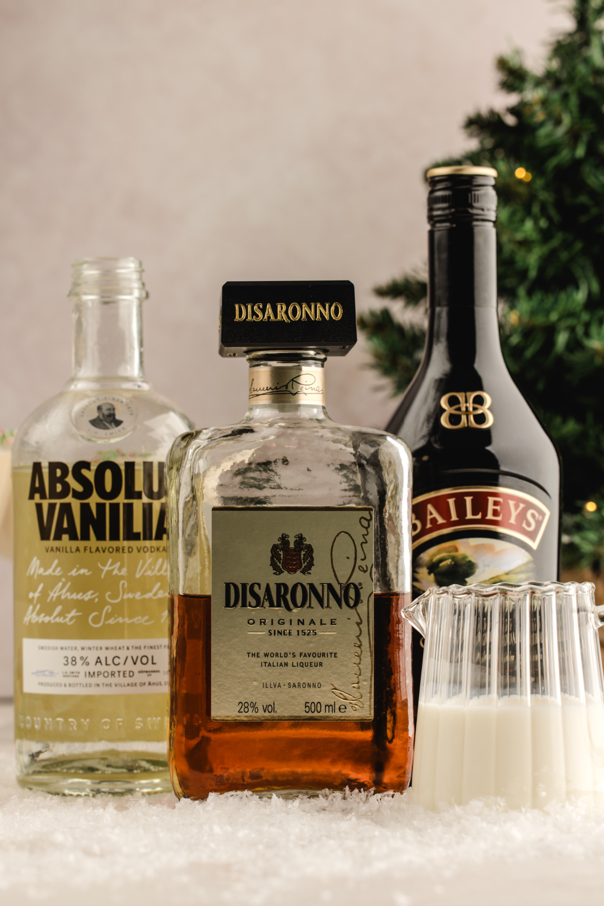 Vanilla vodka, amaretto, Irish cream, and a small pitcher of cream arranged together on a snowy surface with a Christmas tree blurred behind them.