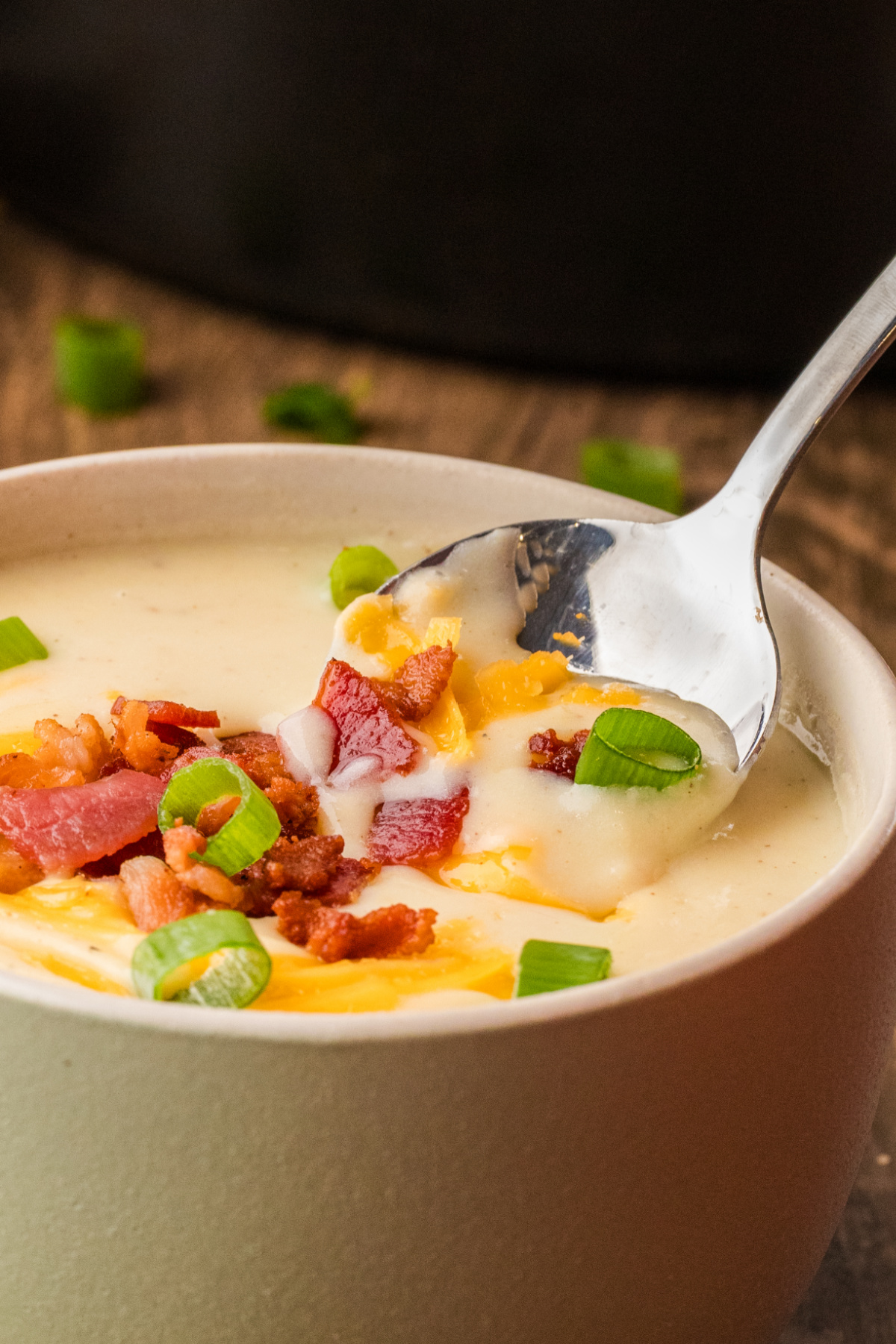 A spoon dipping into loaded baked potato soup, showing chunks of potato, melted cheddar, crisp bacon, and scallions in a creamy base.
