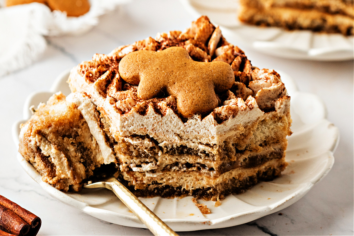 A close-up slice of gingerbread tiramisu dessert on a plate, with a gingerbread cookie on top and a spoonful already taken to show the soft, spiced layers.