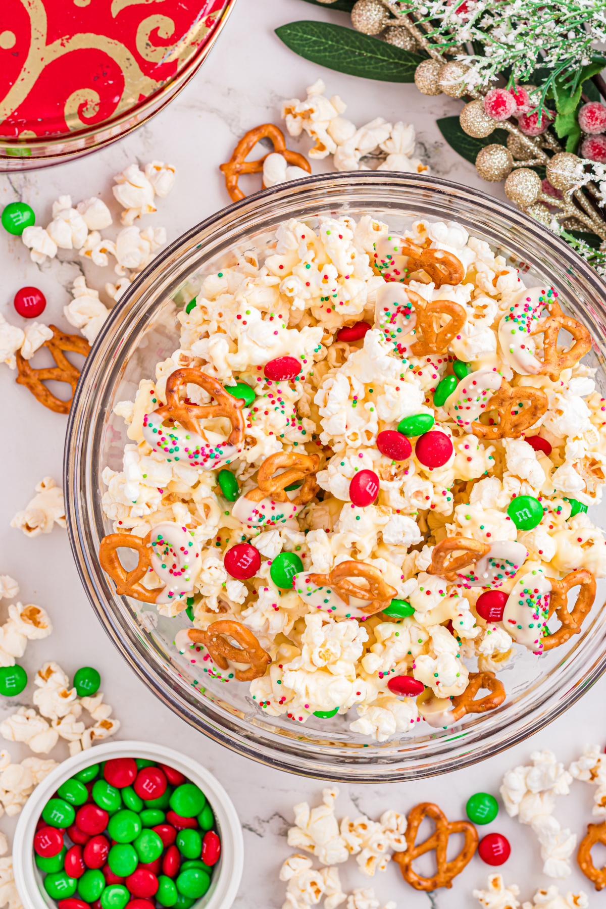 White chocolate popcorn with dipped pretzels, red and green candy-coated chocolates, and holiday sprinkles in a glass bowl.