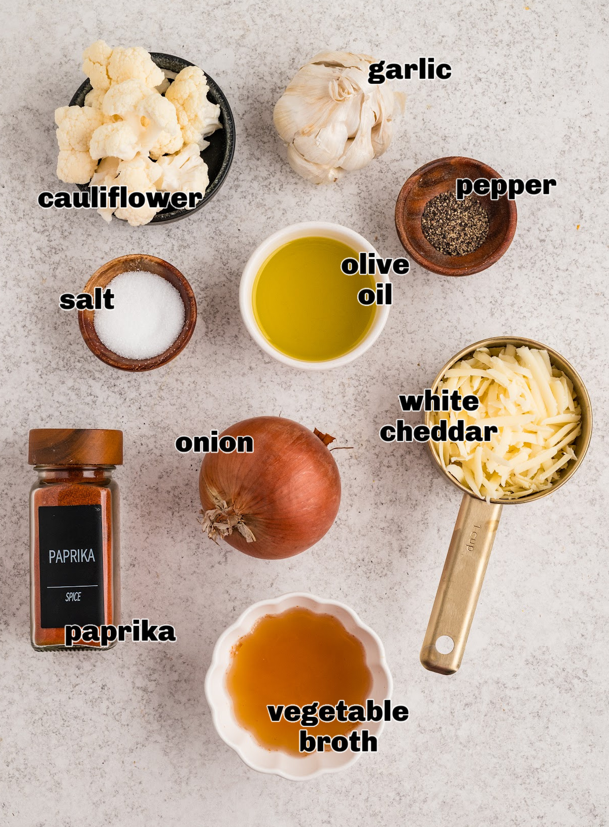 Ingredients for cauliflower soup arranged on a light surface, including cauliflower florets, garlic, onion, olive oil, vegetable broth, white cheddar, salt, pepper, and paprika.