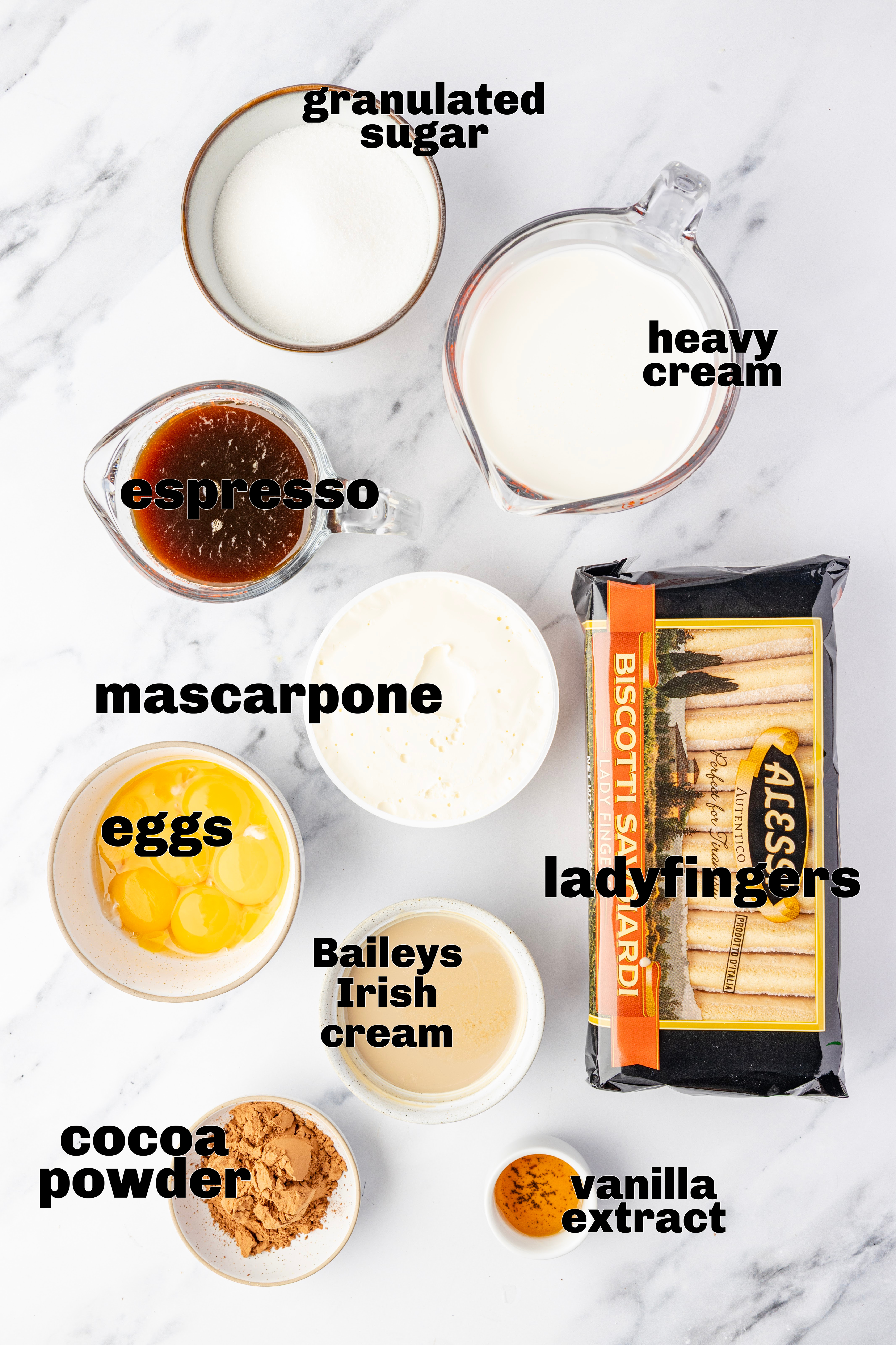 Flat lay of tiramisu ingredients labeled on the image, including mascarpone, eggs, espresso, granulated sugar, heavy cream, ladyfingers, cocoa powder, vanilla extract, and Baileys Irish cream.